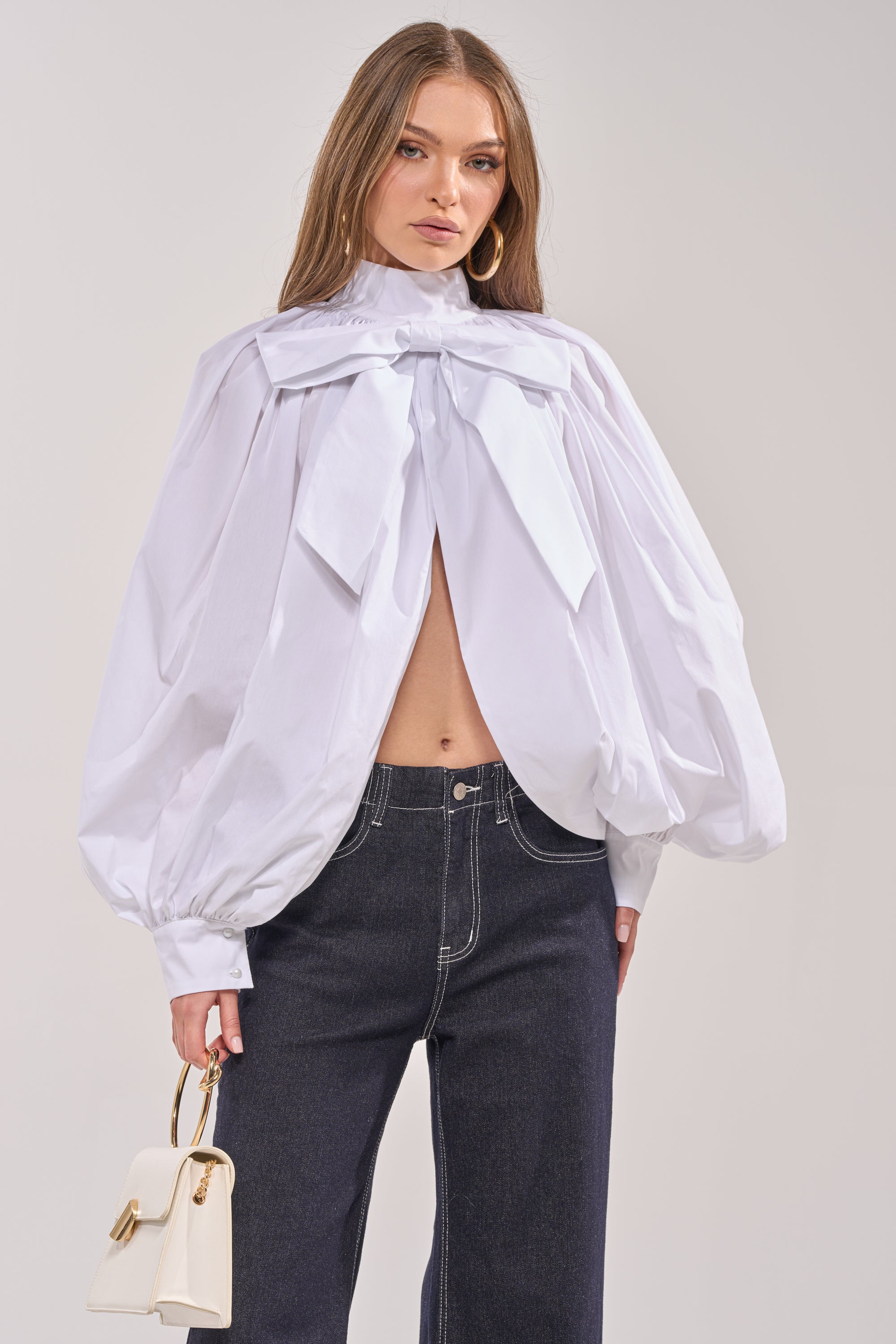 A woman wears the BLACK TIE AFFAIR BLOUSE IN WHITE, featuring an oversized bow-tie neckline and billowy sleeves, paired with dark high-waisted jeans. She accessorizes with a small white handbag and has long, straight hair. 2
