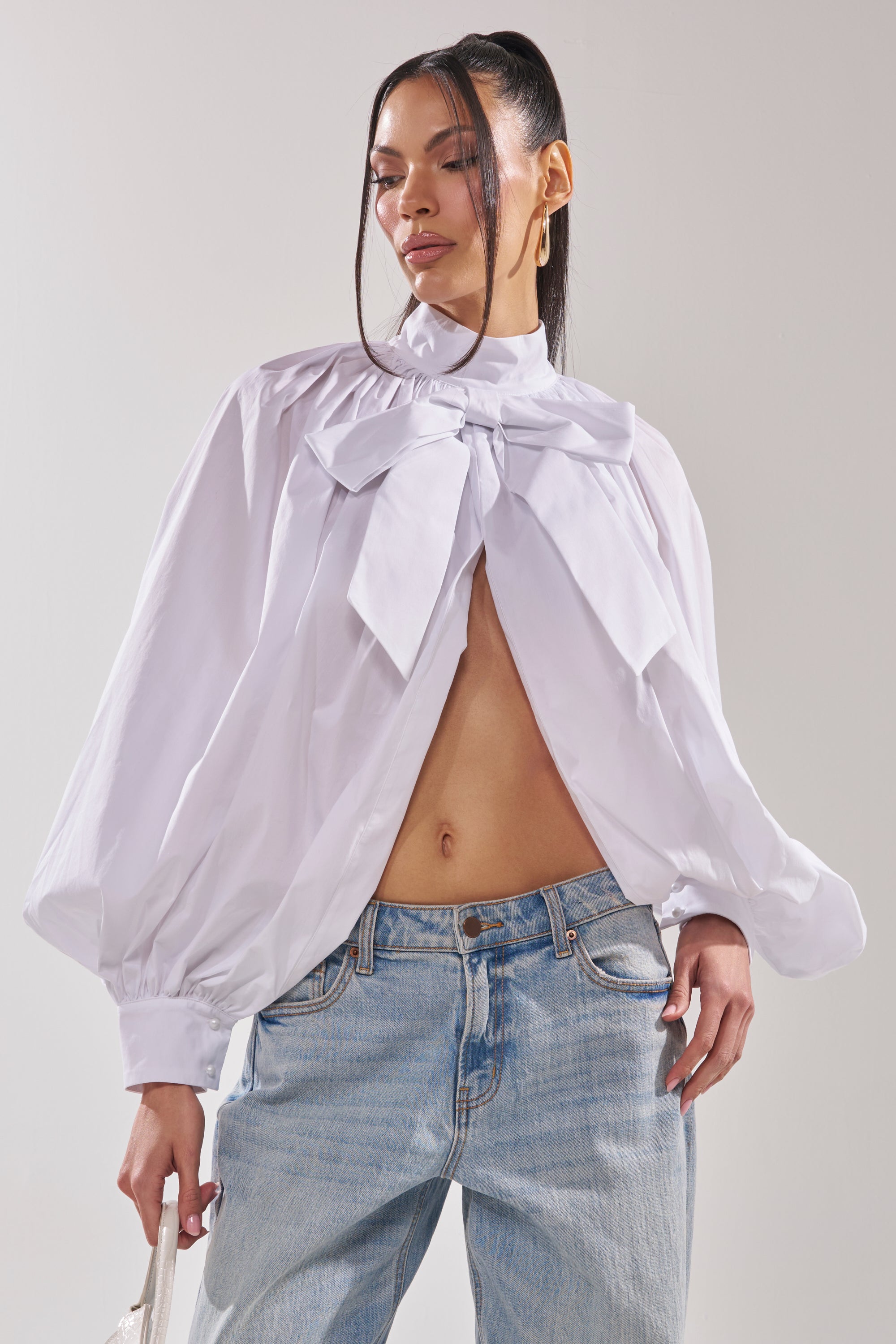A woman wears the BLACK TIE AFFAIR BLOUSE IN WHITE, featuring voluminous long sleeves, a large neckline bow, and an open front that reveals her midriff, paired with light blue jeans. 3