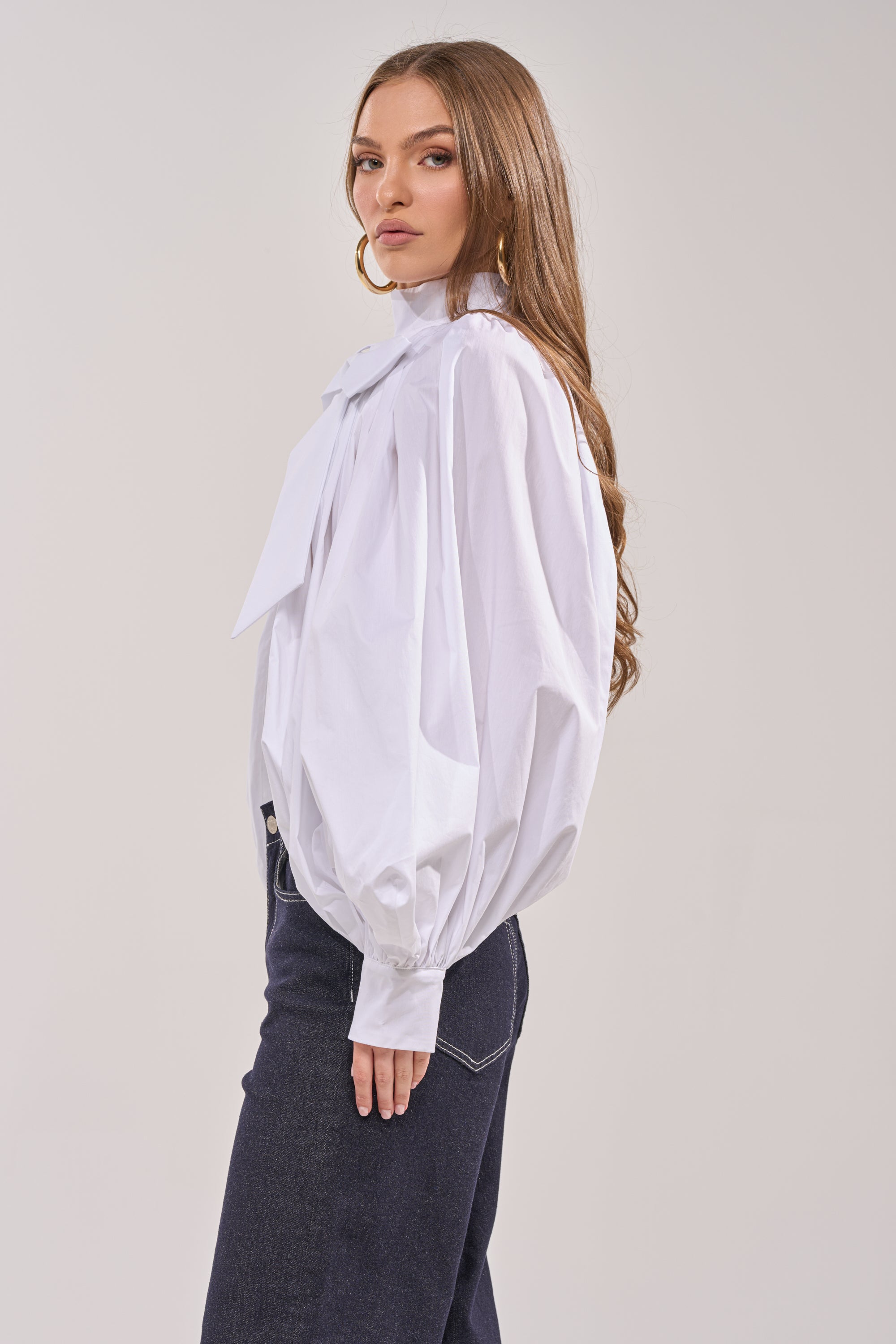 A woman with long brown hair wears the BLACK TIE AFFAIR BLOUSE IN WHITE, featuring an oversized bow-tie neckline and voluminous sleeves, paired with dark jeans. She stands sideways, looking at the camera against a plain light background. 3
