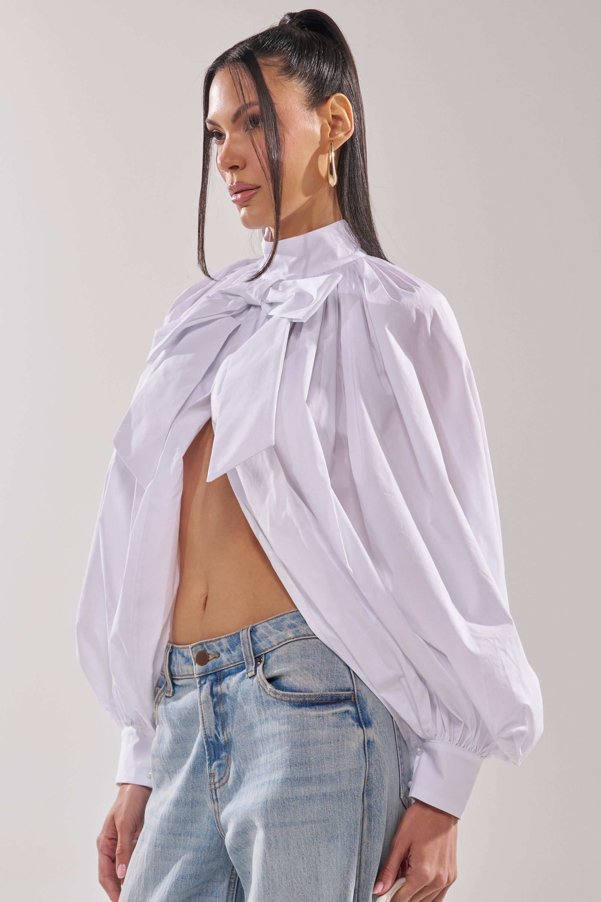 A woman wears the BLACK TIE AFFAIR BLOUSE IN WHITE, featuring oversized bows and balloon sleeves, paired with light blue jeans. Her hair is styled in a high ponytail as she stands against a plain background. 4