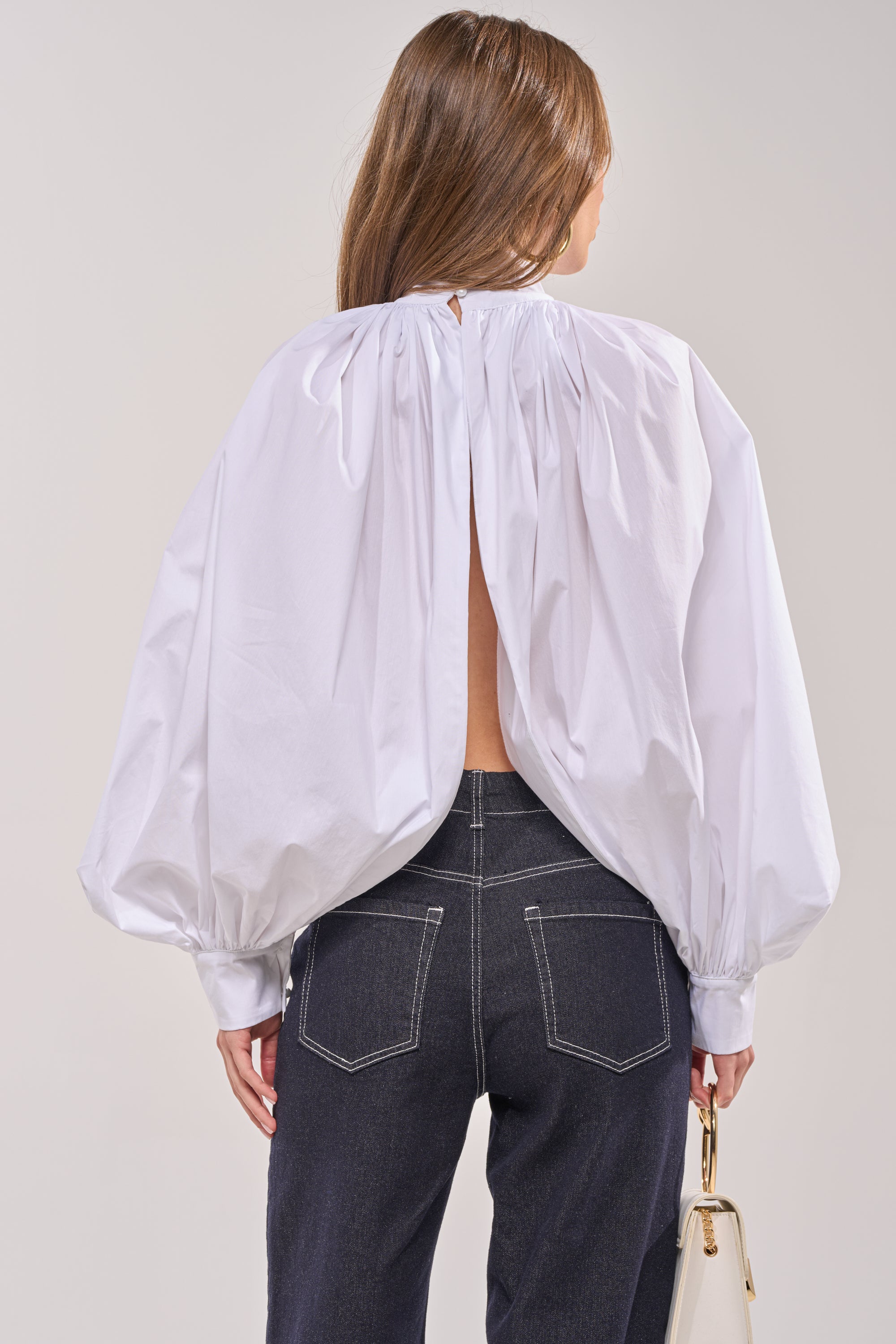 A woman with long brown hair wears the BLACK TIE AFFAIR BLOUSE IN WHITE—featuring voluminous sleeves—with dark high-waisted jeans and a light handbag, seen from behind against a neutral background. 4
