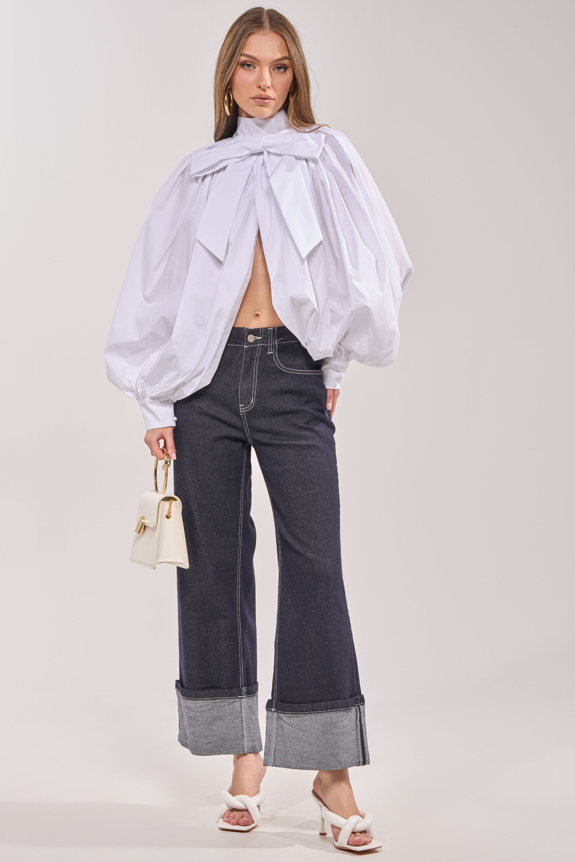A woman wears the BLACK TIE AFFAIR BLOUSE IN WHITE, featuring voluminous long sleeves and a large neck bow, paired with dark wide-leg jeans, white heeled sandals, and a small cream handbag. 5