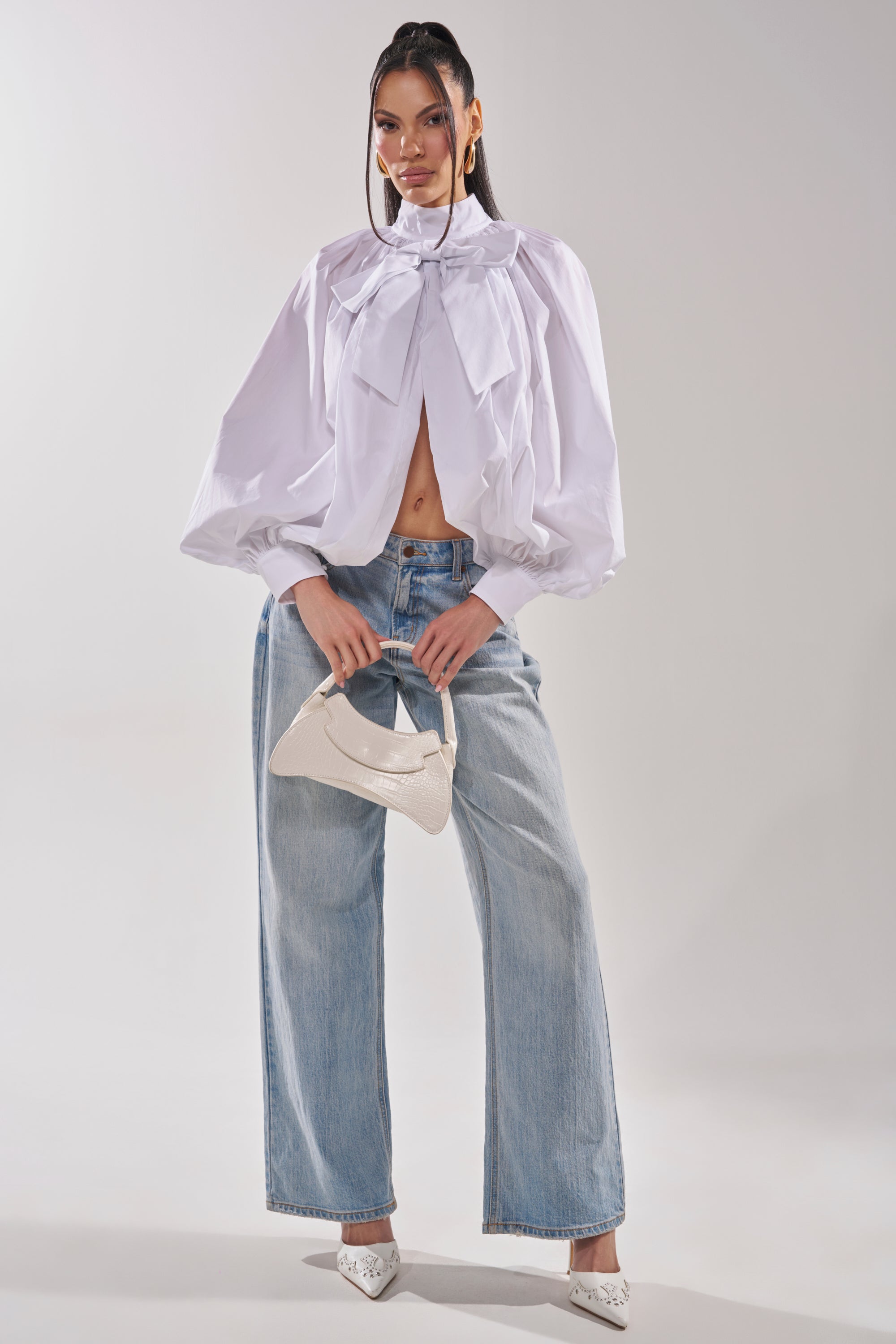 A woman poses in the BLACK TIE AFFAIR BLOUSE IN WHITE with large bow details and puffy sleeves, paired with light blue wide-leg jeans, white pointed heels, and a small white textured handbag against a plain background. 6