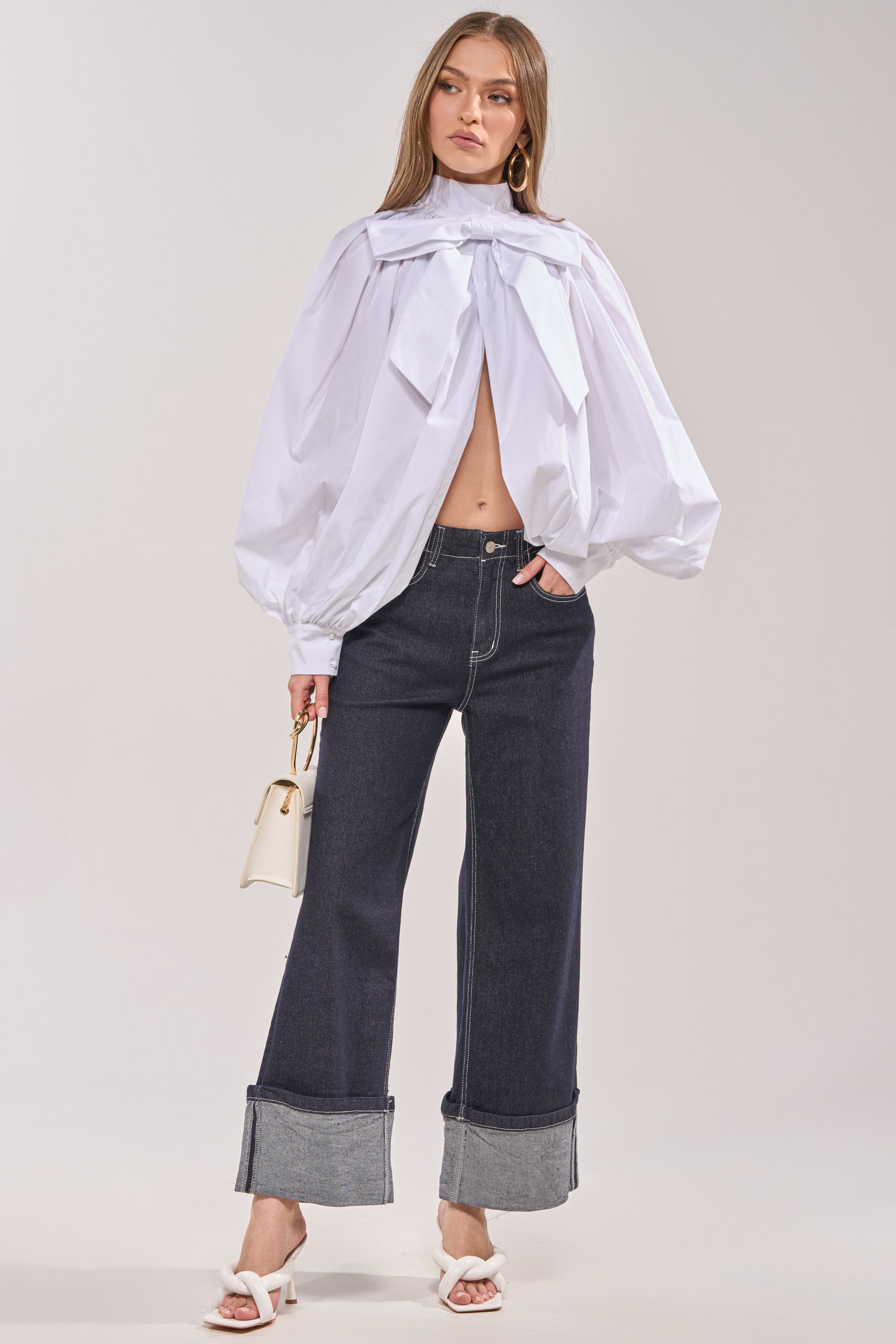 A woman wears the BLACK TIE AFFAIR BLOUSE IN WHITE with dramatic ruffles and a keyhole cutout, styled with high-waisted wide-leg dark jeans with large cuffs, white heeled sandals, and a small white handbag. 6