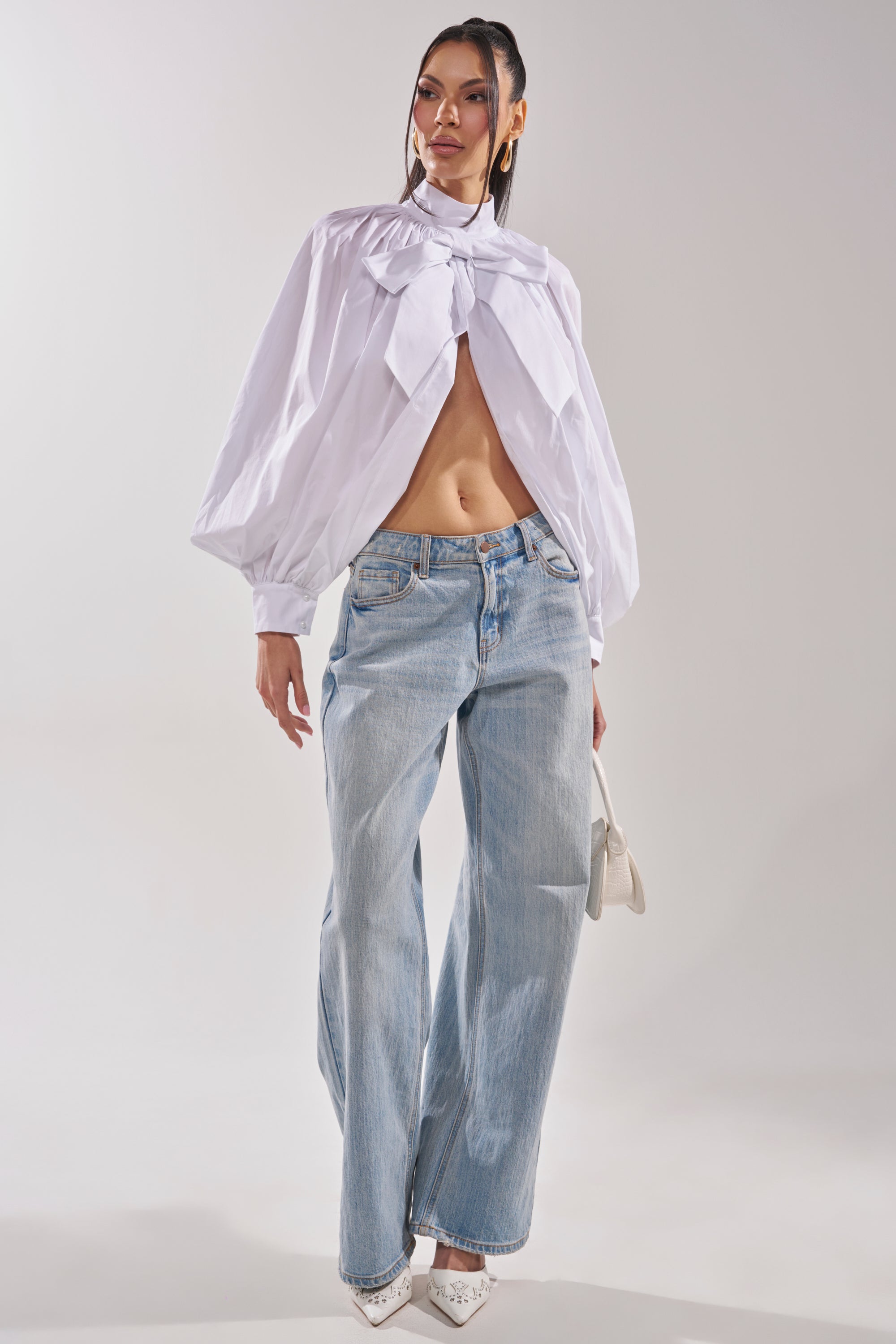 A woman poses in the BLACK TIE AFFAIR BLOUSE IN WHITE with a large bow, paired with light blue wide-leg jeans, white high heels, and a small white handbag against a plain background. 7