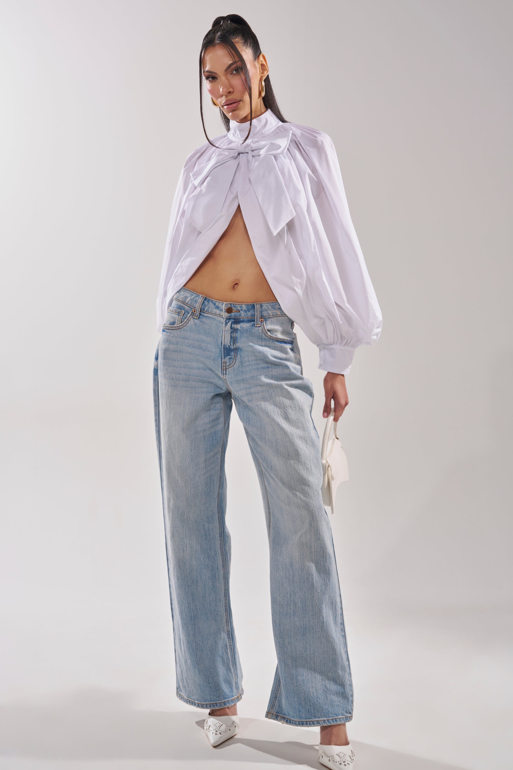 A woman poses confidently in the BLACK TIE AFFAIR BLOUSE IN WHITE, featuring a high neck and bow accent, paired with light blue wide-leg jeans. She accessorizes with a white handbag and pointed-toe heels against a plain backdrop. 8