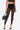 A woman wears SUPER LACE STRETCHY LEGGINGS in sheer black lace with a matching crop top and pointed black heels, holding a shiny black handbag against a plain white background.