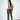 A woman with a high ponytail stands against a white wall, facing away and looking over her shoulder. She wears SUPER LACE STRETCHY LEGGINGS with a high-rise fit, a geometric blue corset, and gold high heels.