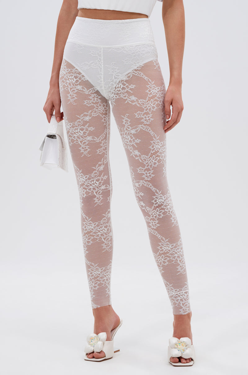 A person models SUPER LACE STRETCHY LEGGINGS in sheer white lace, paired with a white crop top, flower-detailed heeled sandals, and a small white bag against a light background—perfect for flaunting these stylish high-rise leggings.