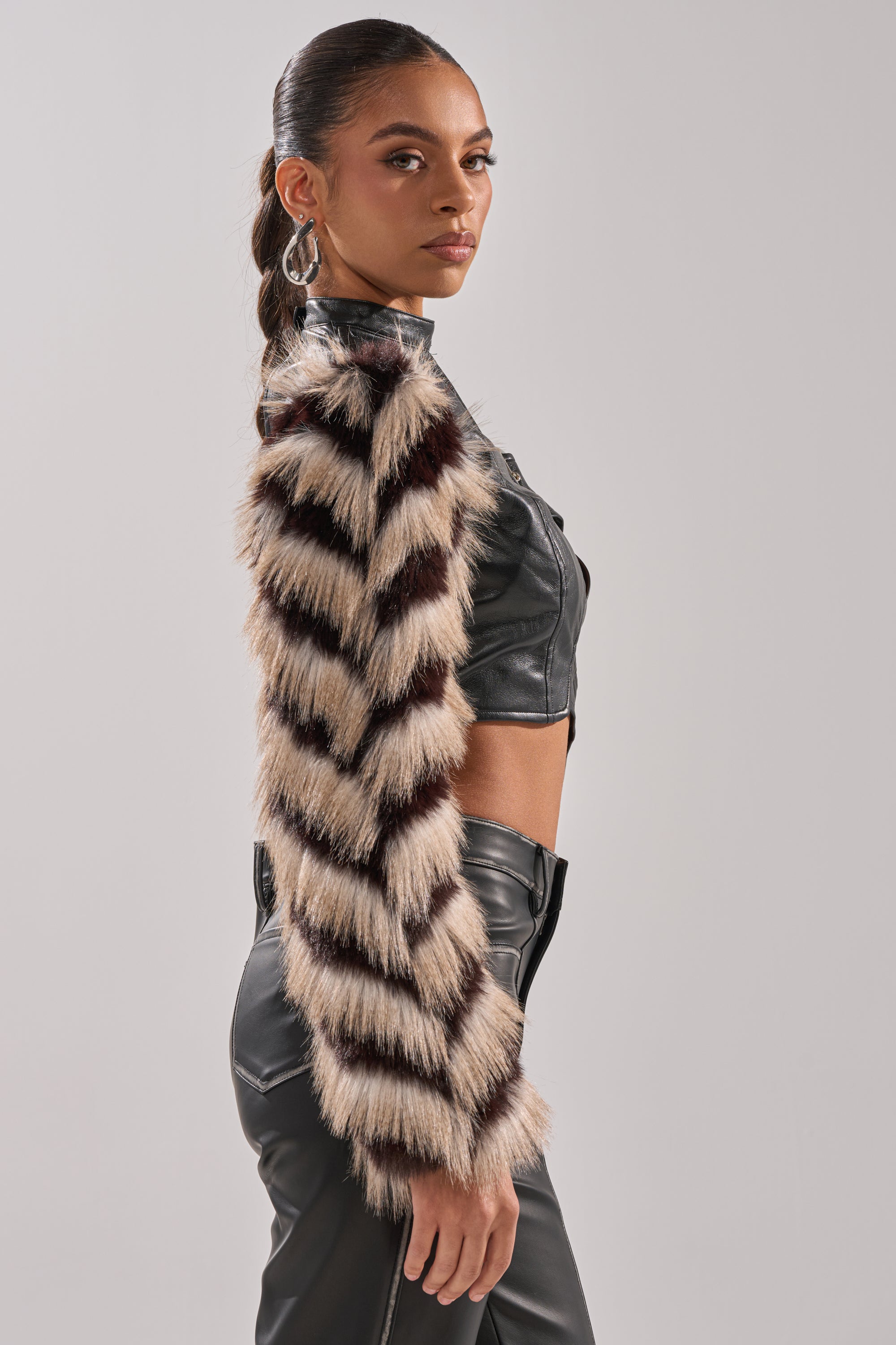 A woman wears the HIGH AND LOW FUR BLOUSE—a furry, striped statement piece—over black leather pants and a cropped faux leather blouse. She poses in profile with a sleek ponytail, confidently glancing over her shoulder against a plain background.