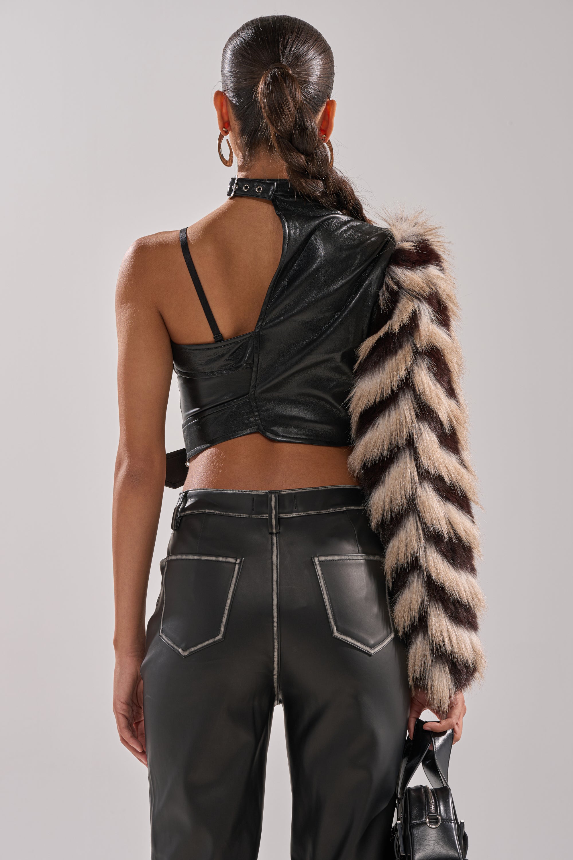 A person with a sleek ponytail holds a small black bag, wearing the HIGH AND LOW FUR BLOUSE—an asymmetrical black faux leather top with one furry striped sleeve—paired with black leather pants featuring white stitching.