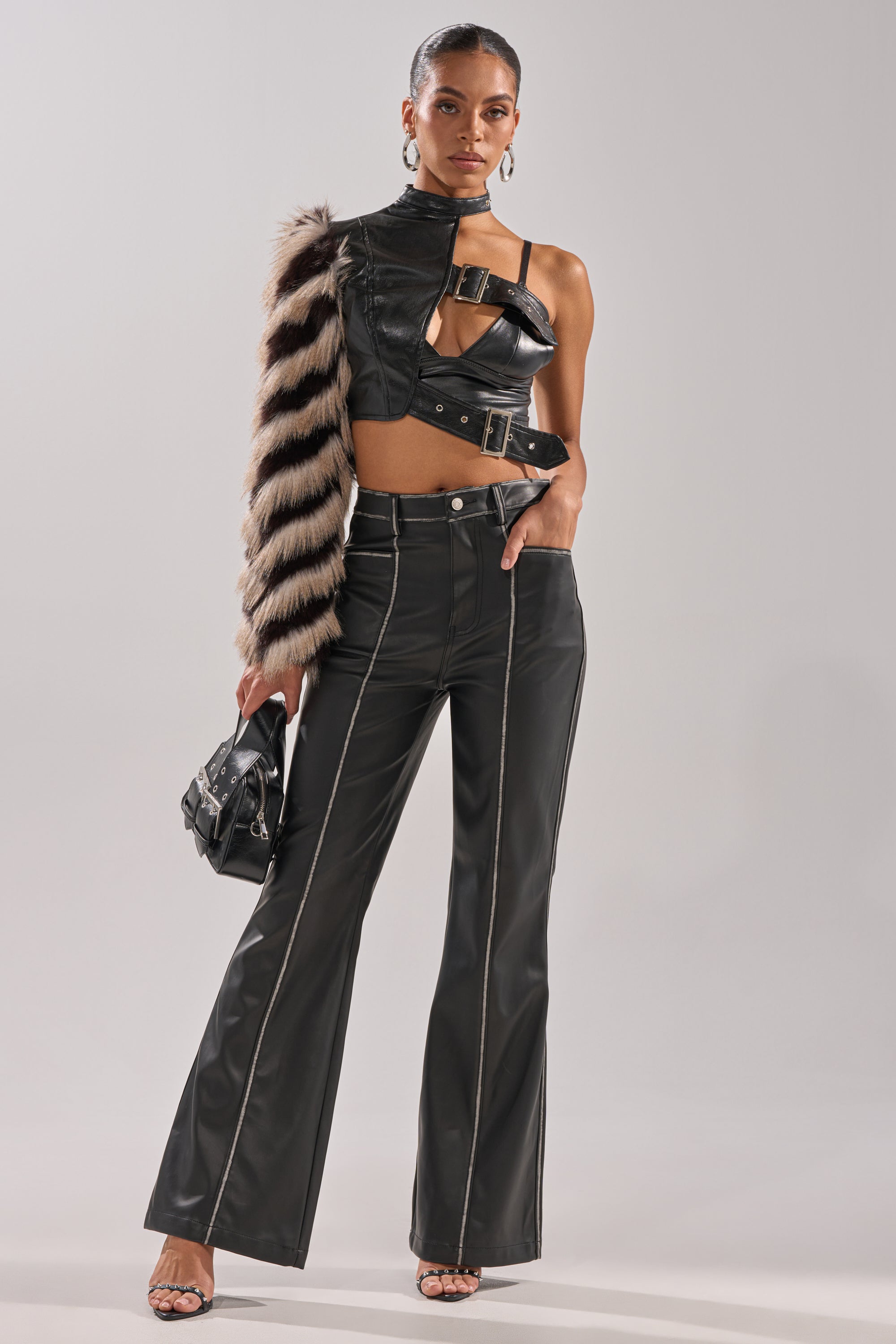 A woman models the HIGH AND LOW FUR BLOUSE, a black faux leather one-shoulder crop top with a chic one-sleeve design, paired with high-waisted black flared pants, a striped fur stole, hoop earrings, strappy heels, and a small black handbag.