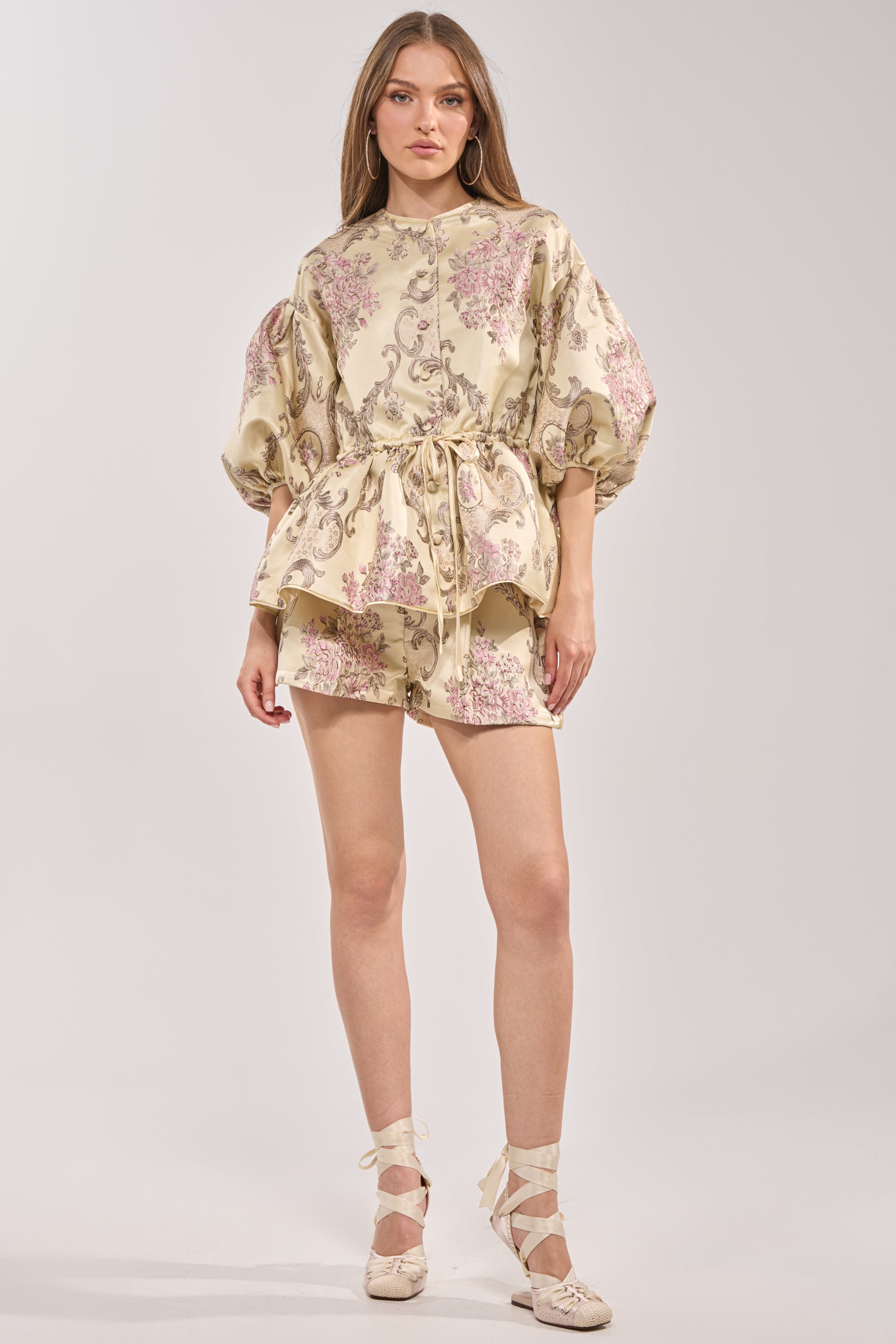 A woman wears the PORCELAIN DOLL BROCADE SHORTS with a matching cream floral top with puffed sleeves, lace-up shoes, and neutral makeup. She poses against a plain background, her long hair styled down. 5