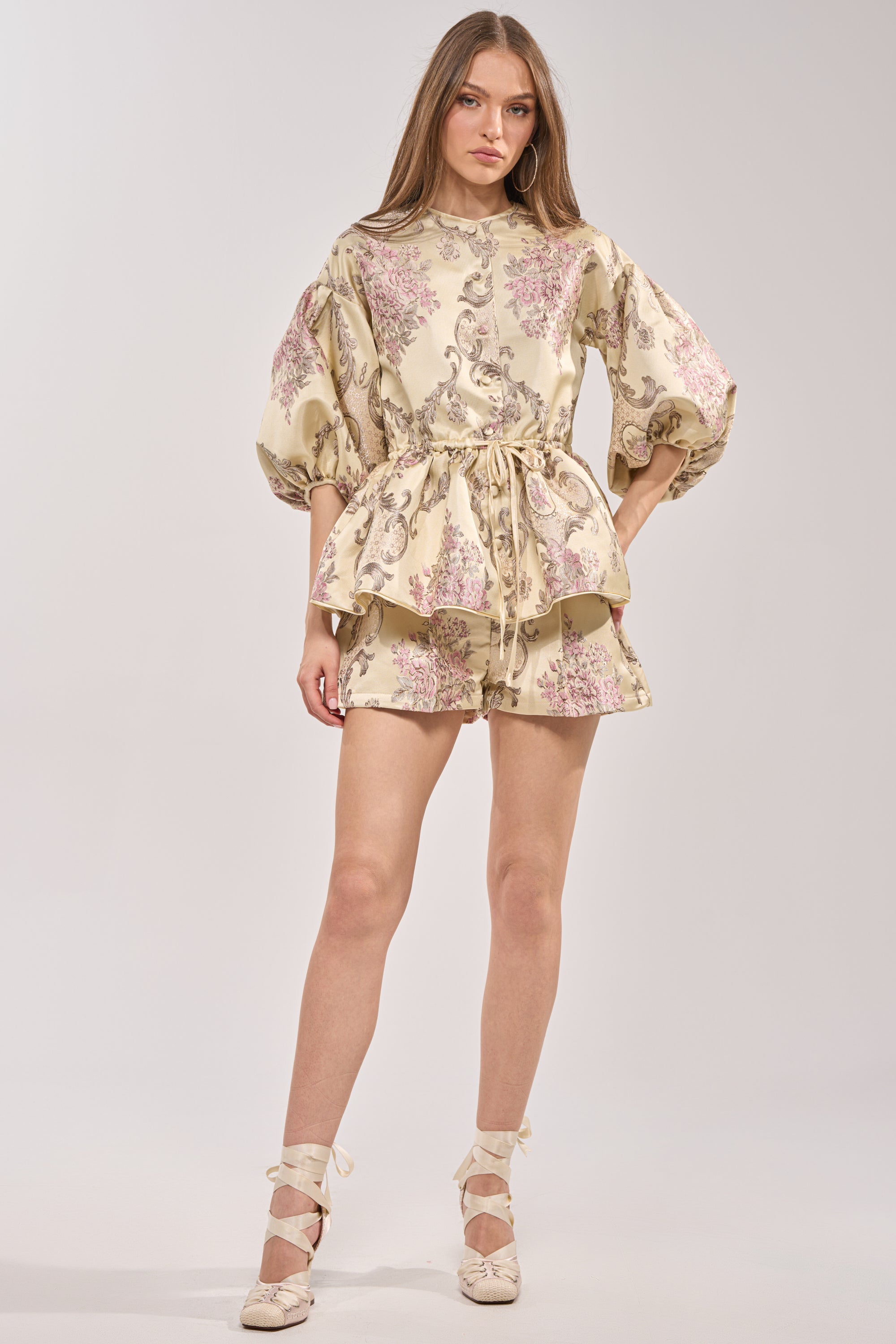 A woman wears the PORCELAIN DOLL BROCADE SHORTS, a high-rise satin style with floral and paisley print, paired with a matching cream top featuring puffy sleeves, a cinched tie waist, lace-up shoes, and loose hair. 6