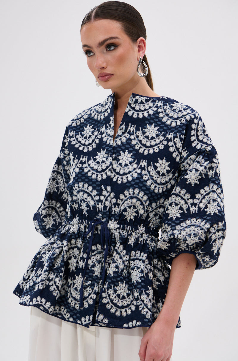 Wearing the MELODY DENIM EYELET BLOUSE in navy with white patterns, a woman with slicked-back hair and large hoop earrings stands against a light background. The cinched waist and long sleeves make this top perfect for summer.