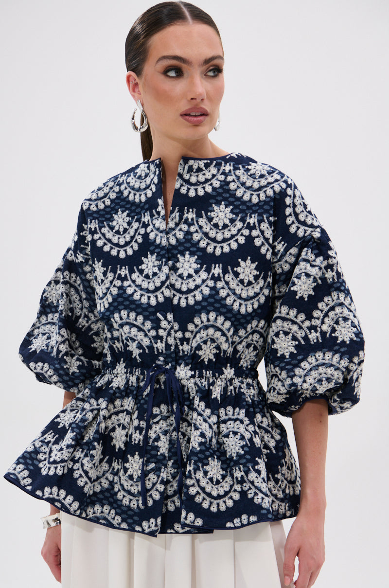 Wearing the MELODY DENIM EYELET BLOUSE in navy with white patterns, puffed sleeves, and a gathered waist, she pairs it with hoop earrings and slicked-back hair for a chic summer look.