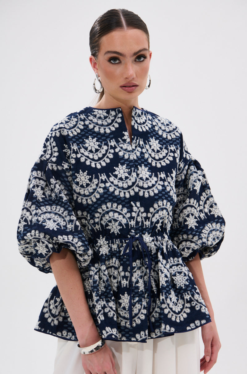 A woman models the MELODY DENIM EYELET BLOUSE—a navy and white statement top with puffed sleeves and a belted waist—styled with white pants, hoop earrings, slicked-back hair, and natural makeup against a plain white background.