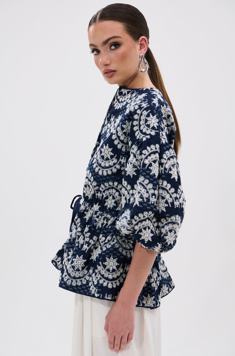 A woman with slicked-back hair models the MELODY DENIM EYELET BLOUSE, a navy blue and white patterned 3/4 sleeve top with a peplum waist, paired with white pants. She stands sideways, gazing at the camera against a plain white background.