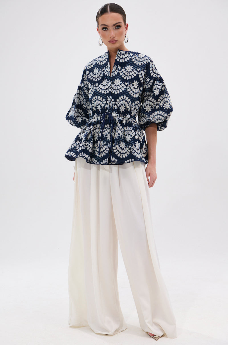 A woman stands against a white background wearing the MELODY DENIM EYELET BLOUSE—navy and white floral, puffed sleeves, cinched waist—paired with high-waisted wide-leg white trousers and large hoop earrings.