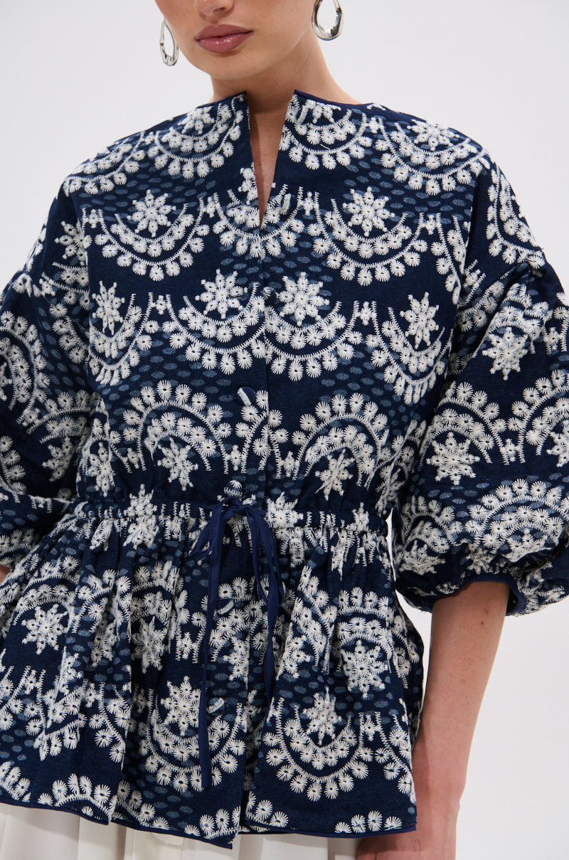 Wearing the MELODY DENIM EYELET BLOUSE—a navy top with white embroidery, split neckline, puffed sleeves, and waist tie—a woman is shown from the waist up, making a perfect summer style statement.
