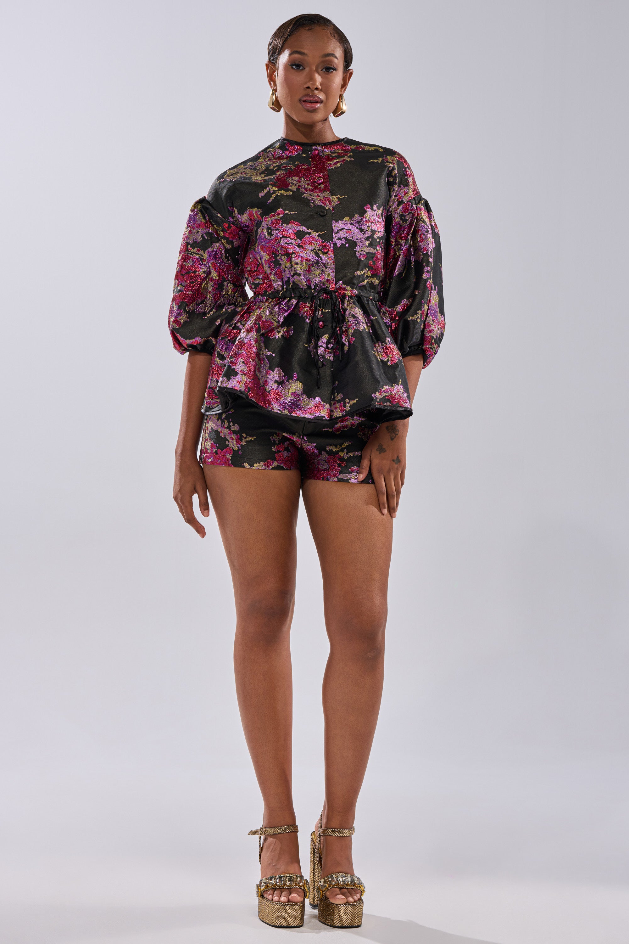 A woman poses confidently against a plain light background, wearing a black puff-sleeve top, ETHEREAL BROCADE SHORTS IN PINK, and gold platform heels.