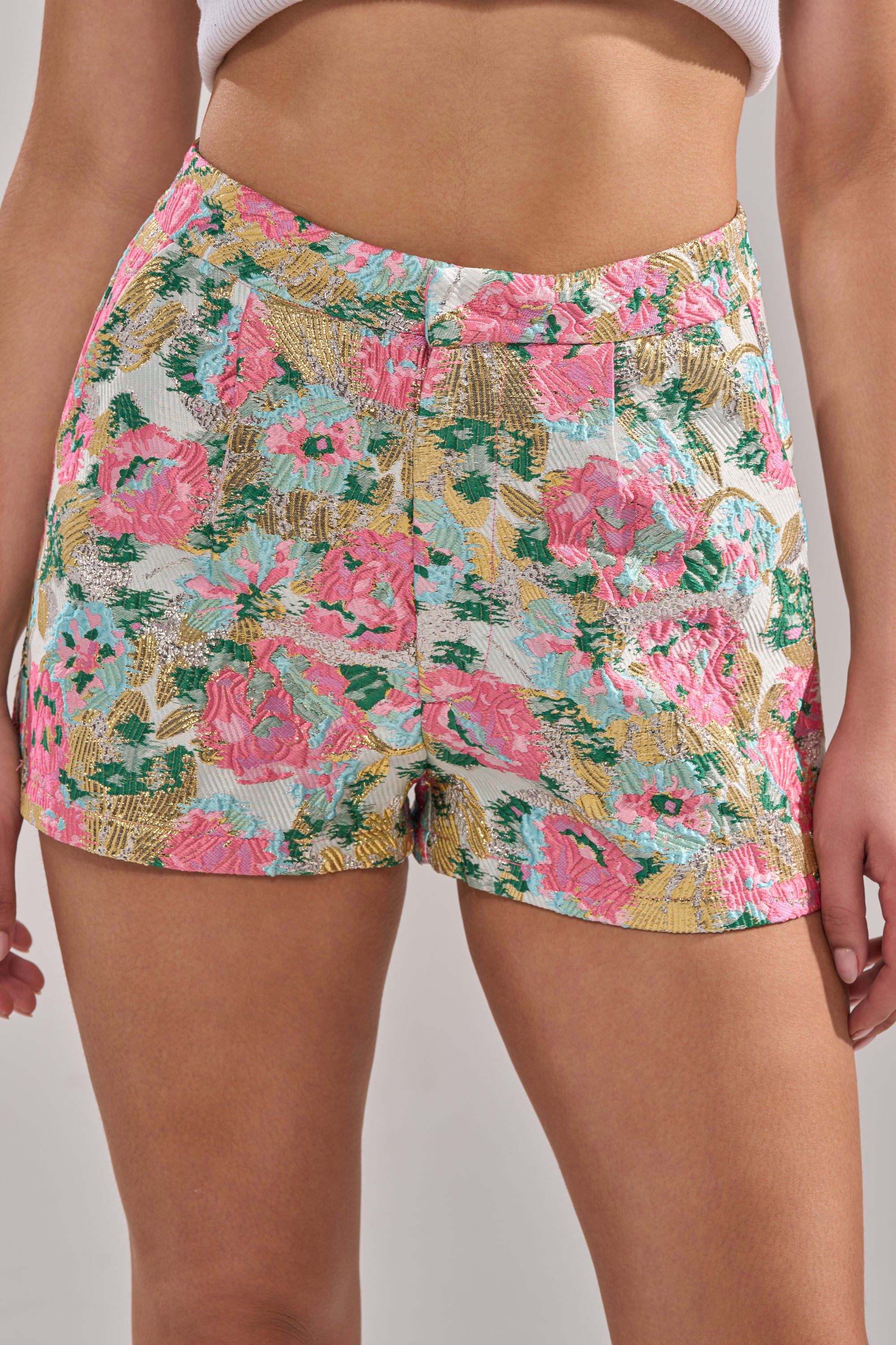 Wearing the TEA TIME FLORAL BROCADE SHORT—high-waisted with vibrant pink, green, yellow, and white floral patterns—paired with a white cropped top. The focus is on the shorts and midsection, hands resting by the sides. 0
