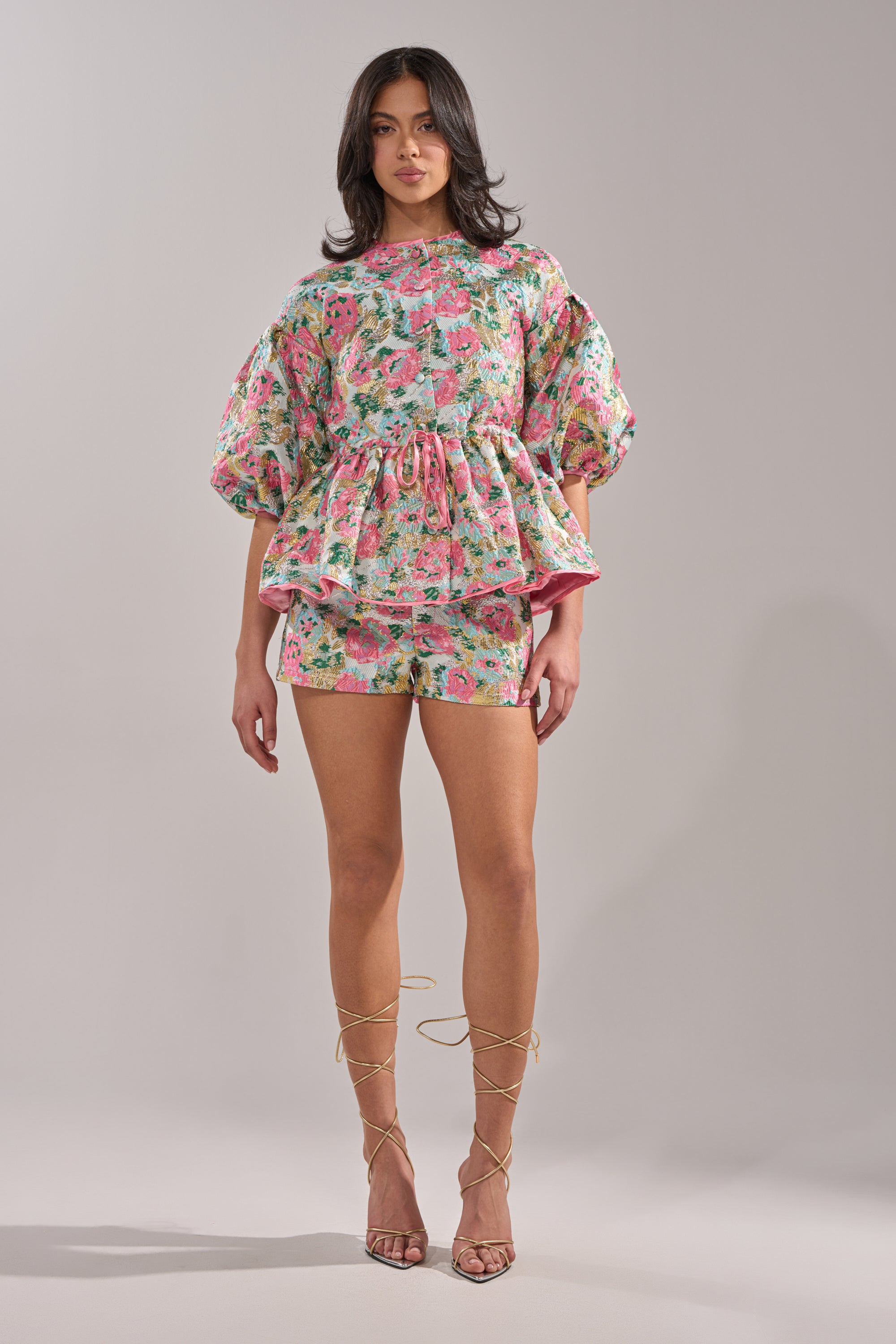 A woman wears the TEA TIME FLORAL BROCADE SHORT with a colorful floral peplum top and puffed sleeves, pairing the look with strappy gold high-heeled sandals against a plain light gray background. 6