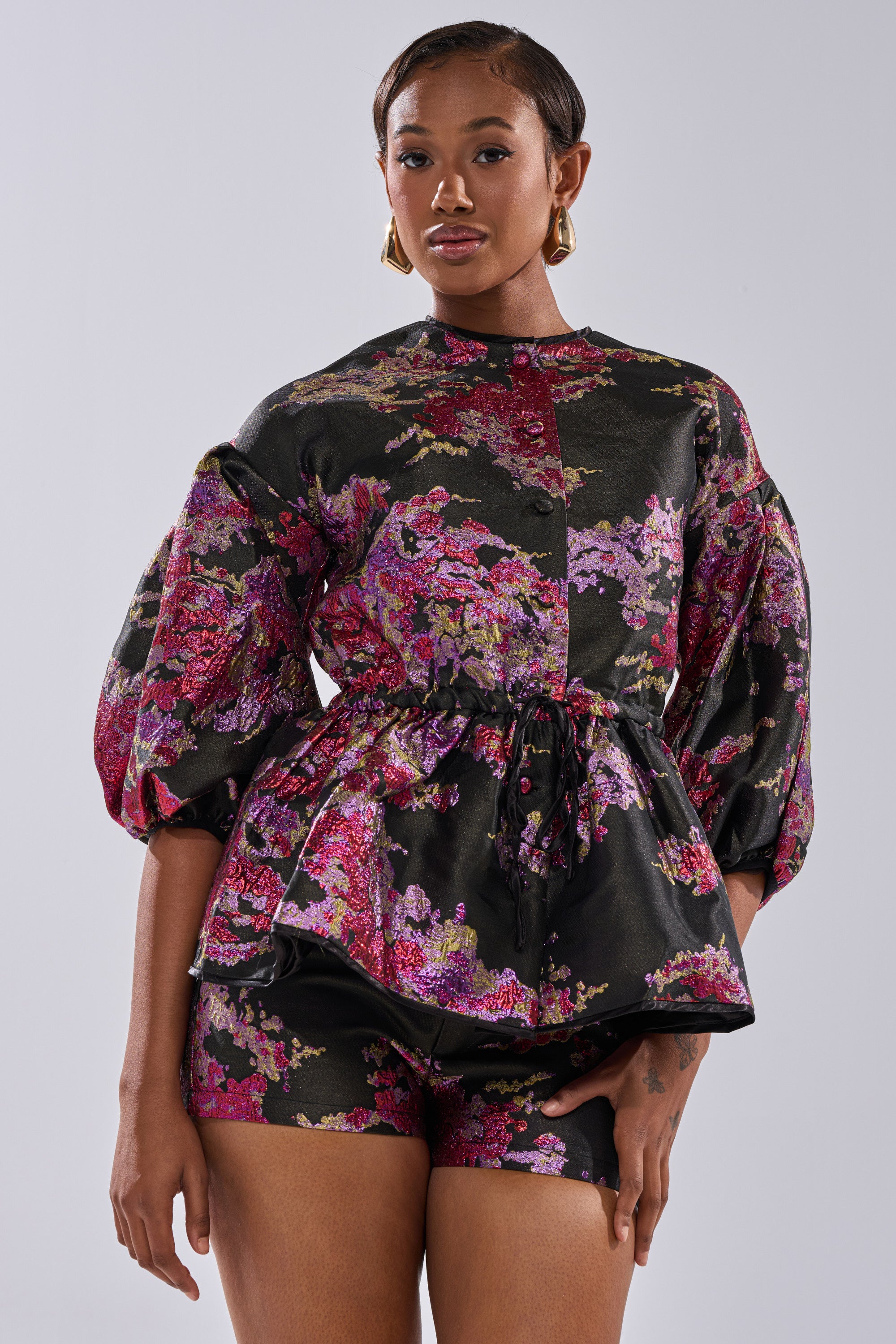 A woman wears the ETHEREAL BROCADE SHORT SLEEVE BLOUSE IN PINK, featuring voluminous sleeves and a vintage floral design. She pairs it with matching shorts and gold earrings, her short hair styled as she poses against a plain background.