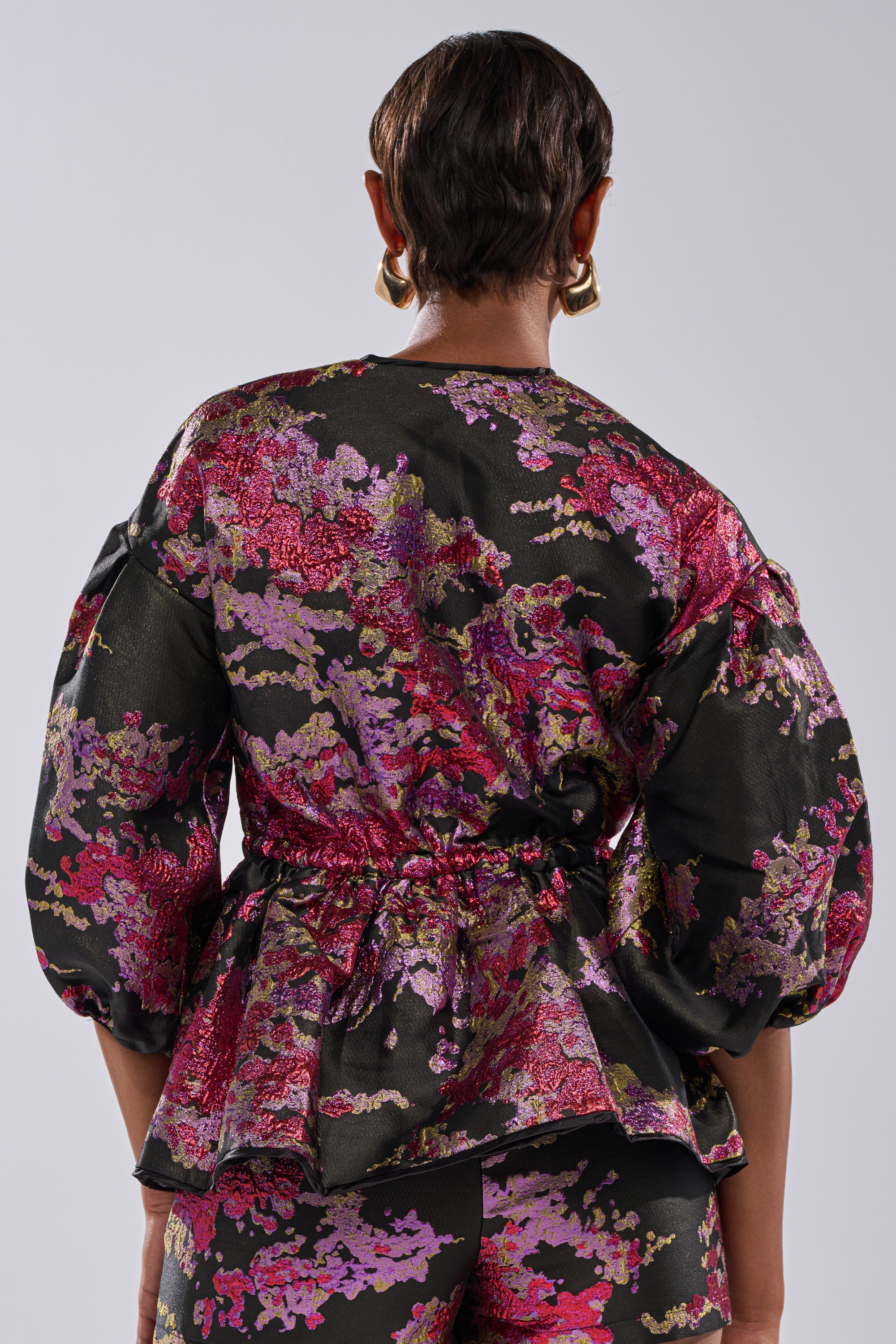 A person with short hair is seen from the back wearing the ETHEREAL BROCADE SHORT SLEEVE BLOUSE IN PINK, paired with matching pants and a cinched waist.