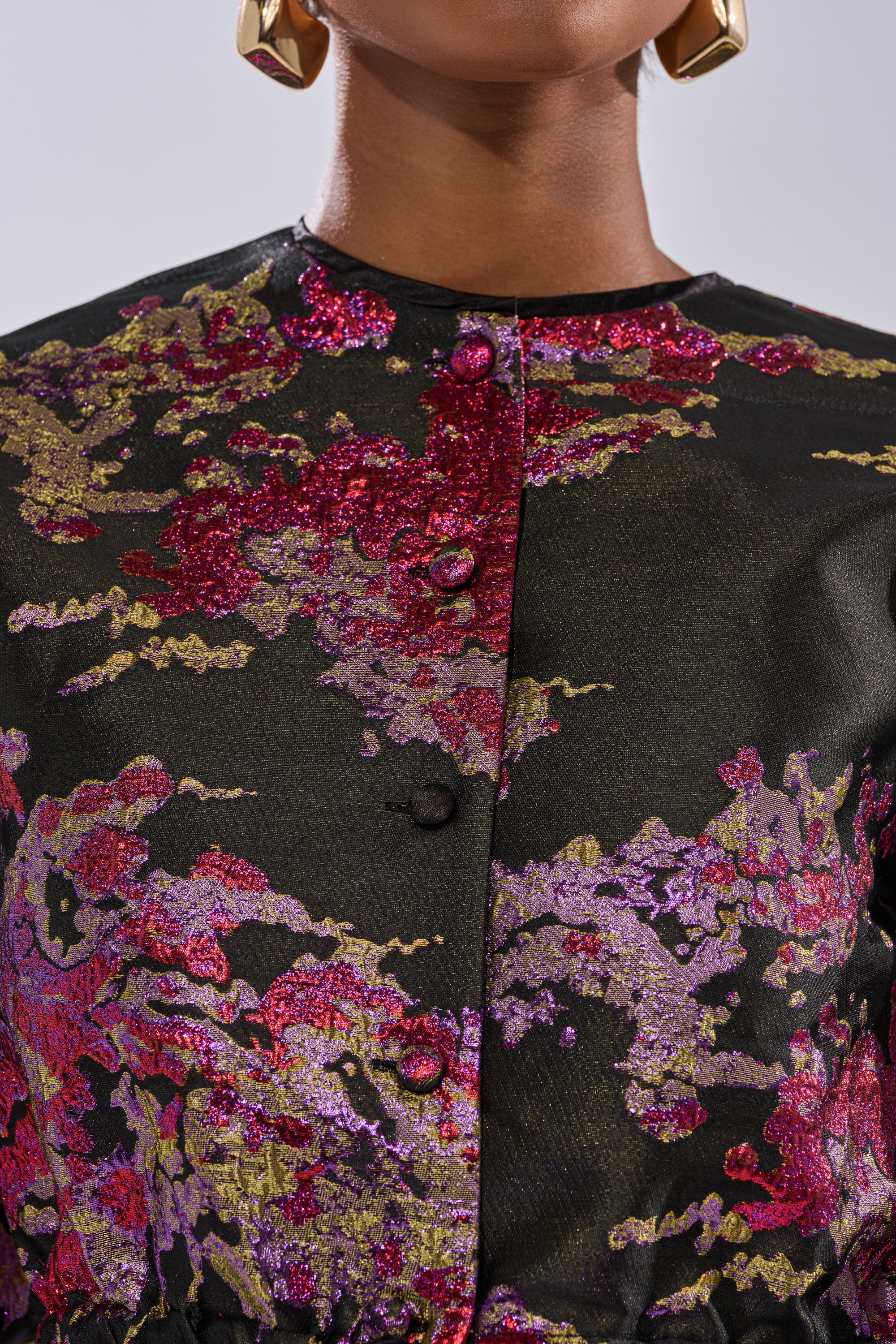A close-up of a person wearing the Ethereal Brocade Short Sleeve Blouse in Pink—a black buttoned shirt with a metallic floral pattern in pink, purple, and gold—paired with large geometric gold earrings.