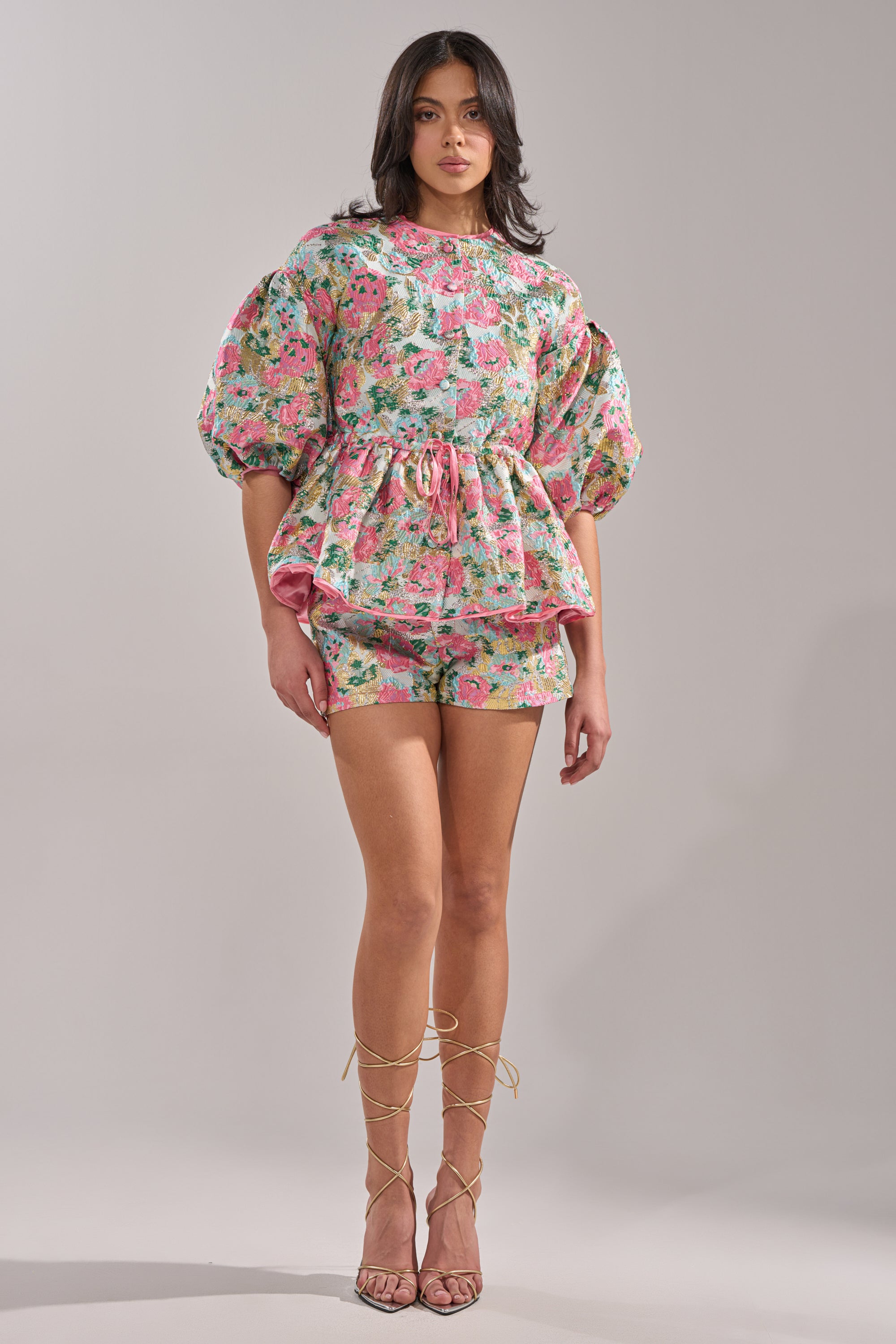 A woman poses in a studio wearing the TEA TIME FLORAL BROCADE SHORT SLEEVE BLOUSE with matching shorts and strappy sandals against a plain, neutral background.