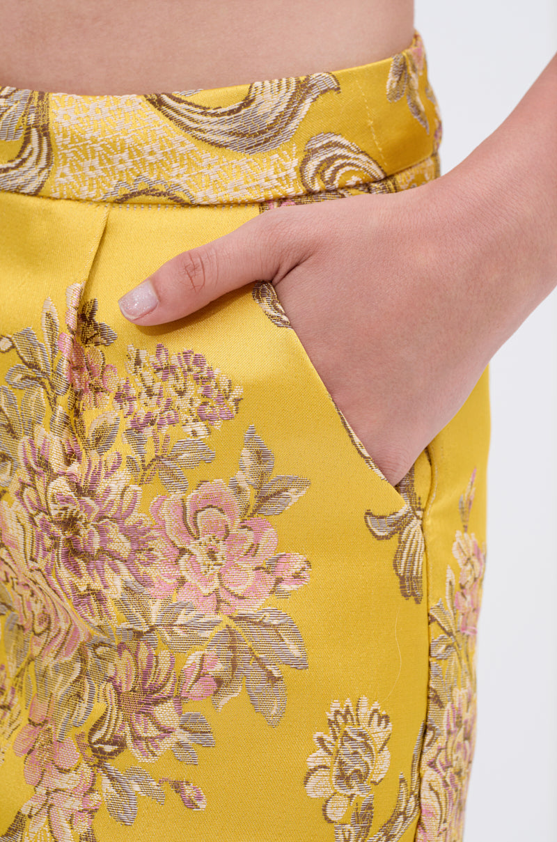 A close-up shows a hand in the pocket of ETHEREAL BROCADE SHORTS, featuring a yellow satin brocade with pink, green, and gold floral patterns. The waistband and top of the shorts are also visible.