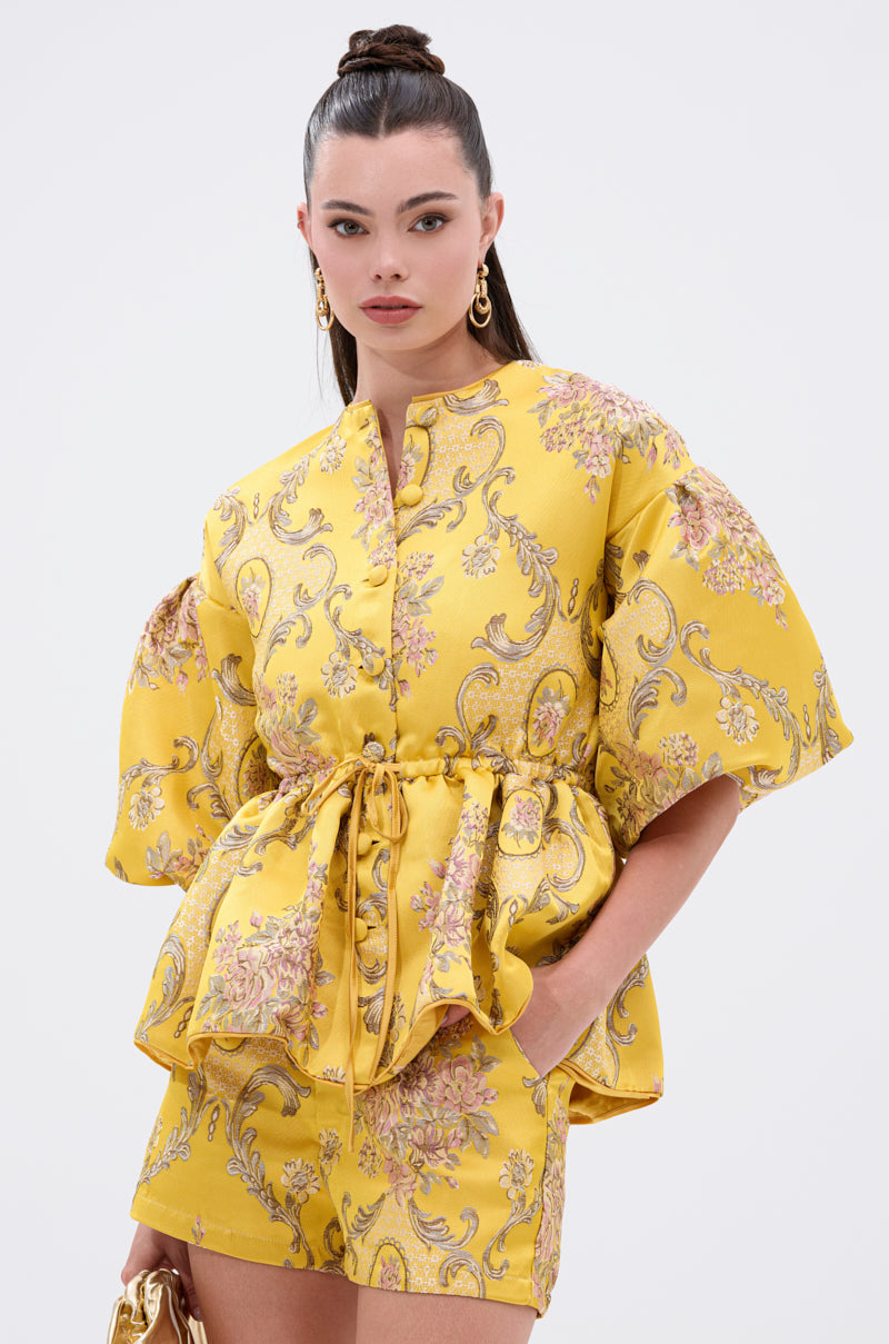 A woman stands against a plain white background wearing the ETHEREAL BROCADE SHORT SLEEVE BLOUSE with puffed sleeves, paired with gold hoop earrings and an updo hairstyle.