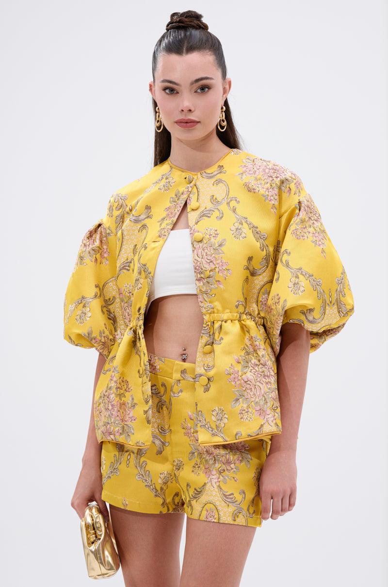 A woman wears the ETHEREAL BROCADE SHORT SLEEVE BLOUSE in a yellow, vintage-inspired floral print with voluminous sleeves, paired with matching shorts and a white crop top. She accessorizes with a small gold purse, gold earrings, and a high ponytail.