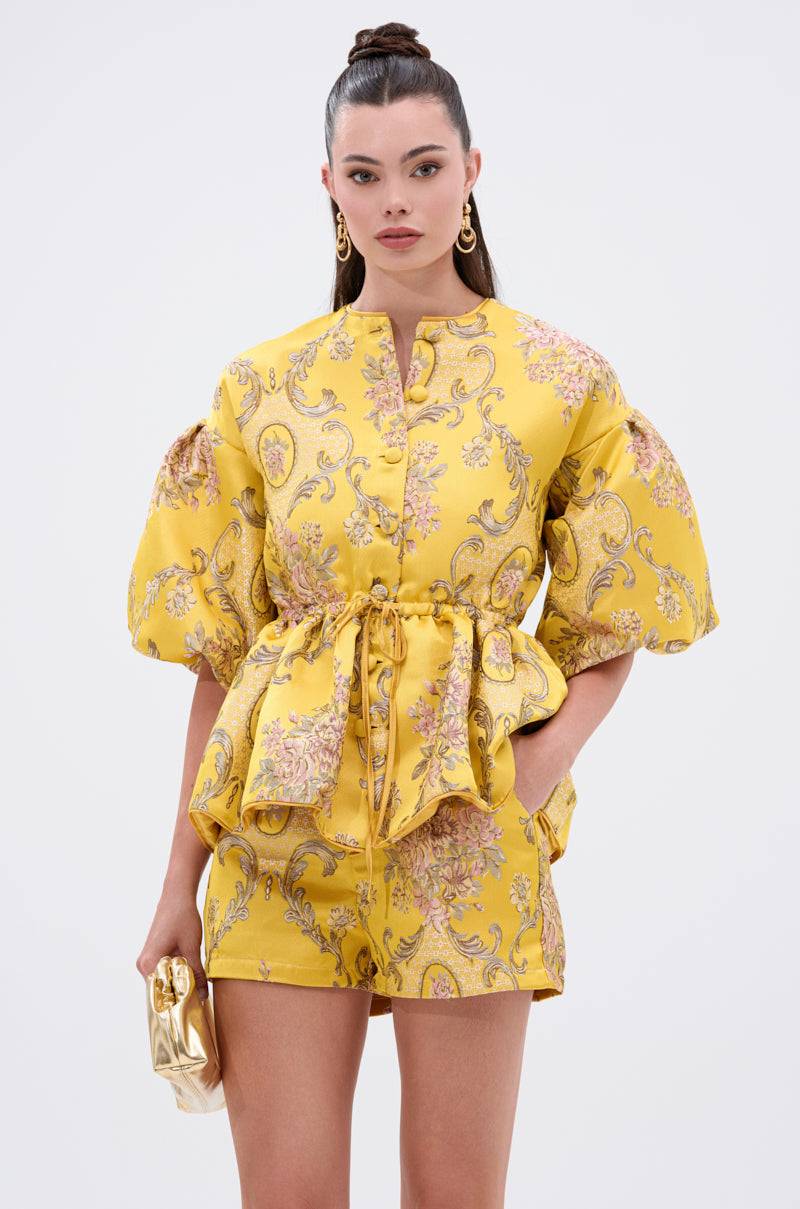 A woman stands against a plain background wearing the ETHEREAL BROCADE SHORT SLEEVE BLOUSE with shorts. The vintage-inspired floral blouse has puffed sleeves. She holds a gold clutch, her hair in a topknot, and wears gold hoop earrings.