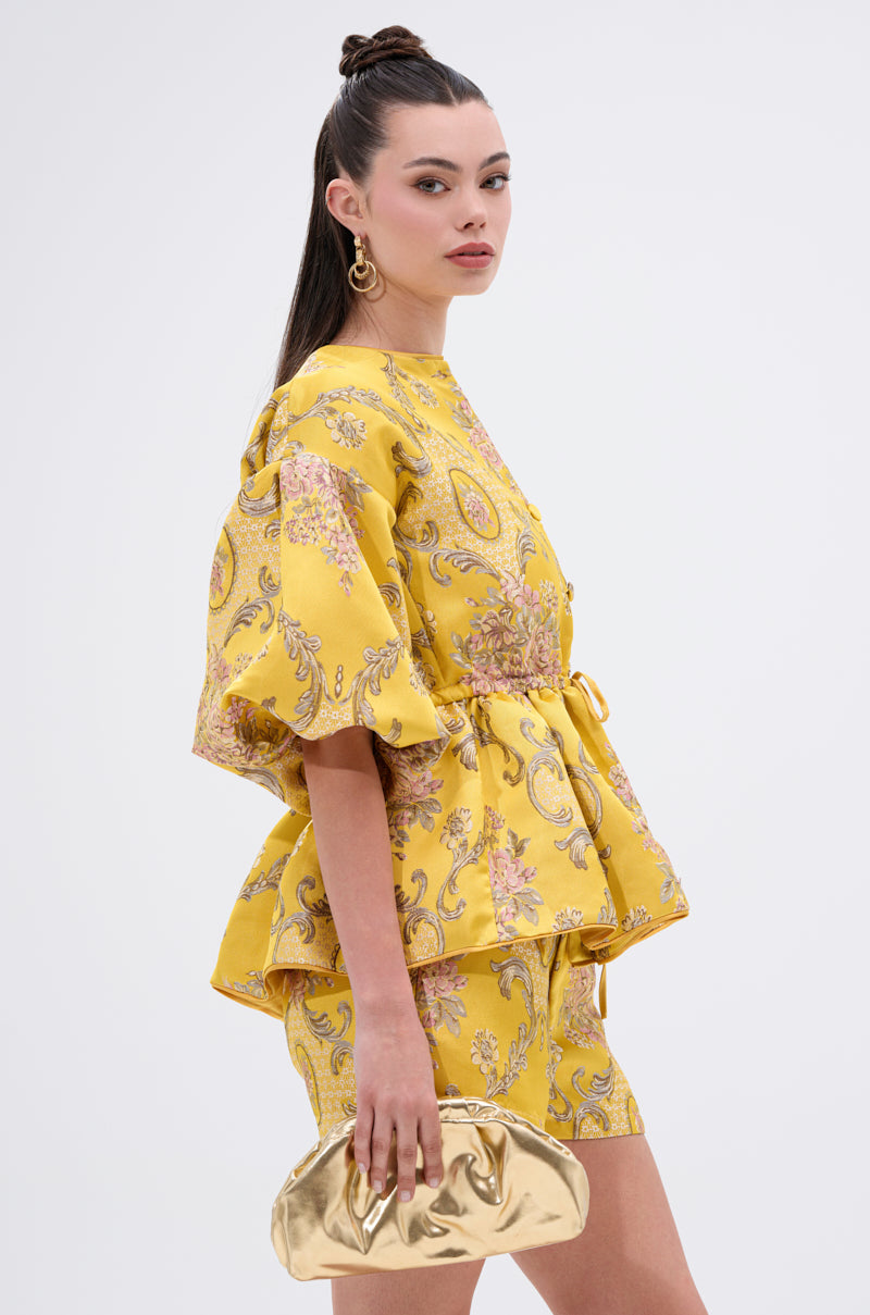 A woman stands against a plain white background wearing the ETHEREAL BROCADE SHORT SLEEVE BLOUSE in a vintage-inspired yellow floral pattern with matching shorts, gold hoop earrings, and a shiny gold clutch. Her long hair is half-up styled.