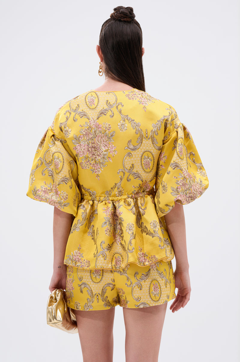 A person with long brown hair in a top knot is seen from behind, wearing the ETHEREAL BROCADE SHORT SLEEVE BLOUSE and matching floral shorts, holding a small gold handbag against a plain white background.