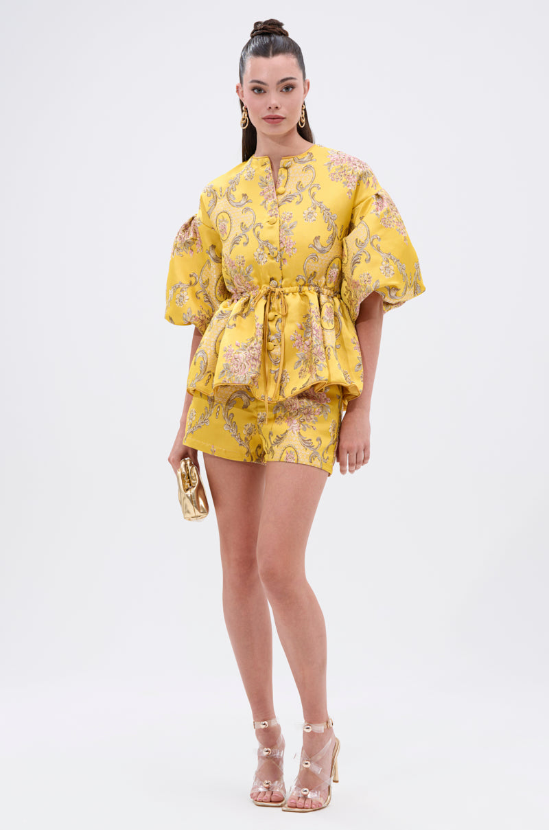 A woman wears the ETHEREAL BROCADE SHORT SLEEVE BLOUSE with a vintage floral pattern, matching shorts, gold earrings, a gold clutch, and clear high-heeled sandals against a plain background.