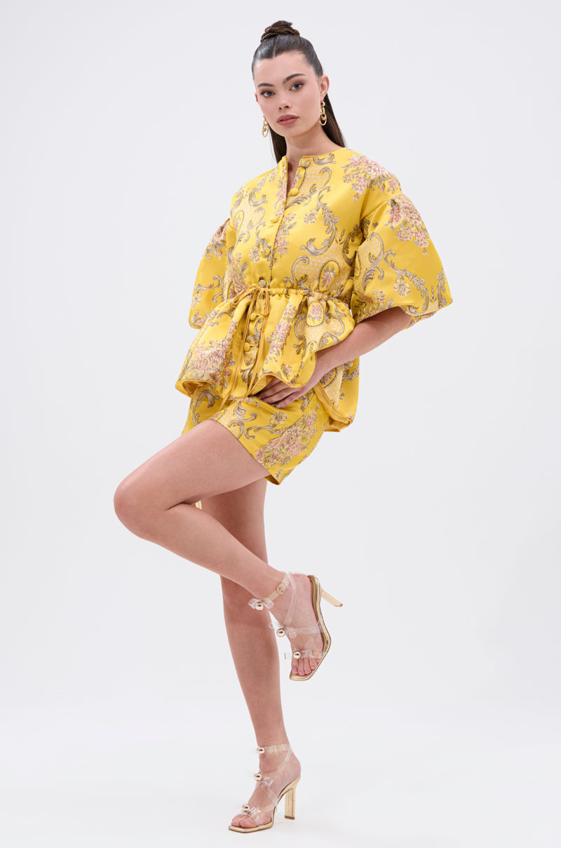 A woman models the ETHEREAL BROCADE SHORT SLEEVE BLOUSE with a vintage-inspired yellow floral pattern and puffed sleeves, styled with a waist belt, clear high-heeled sandals, and gold earrings against a plain white background.