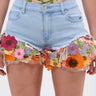 Wearing the VIVI FLORAL DENIM SHORT—high-rise denim adorned with colorful floral embroidery—and a sheer top with matching appliques creates a vibrant, playful look with orange, pink, purple, and yellow blooms and green leaves.