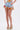Wearing the VIVI FLORAL DENIM SHORT with embroidered floral appliques and tulle ruffles, a sheer flower-pattern top, and straw rainbow-striped platform heels, a person poses against a plain white background.