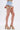 Wearing the VIVI FLORAL DENIM SHORT with embroidered floral appliques and tulle, paired with platform heels featuring woven texture and rainbow-striped soles, a person stands against a plain white background.