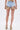 A person wearing the VIVI FLORAL DENIM SHORT—a light blue mini skirt with floral embroidery and a tulle ruffle hem—paired with rainbow-striped high-heeled sandals, seen from the back against a plain white background.