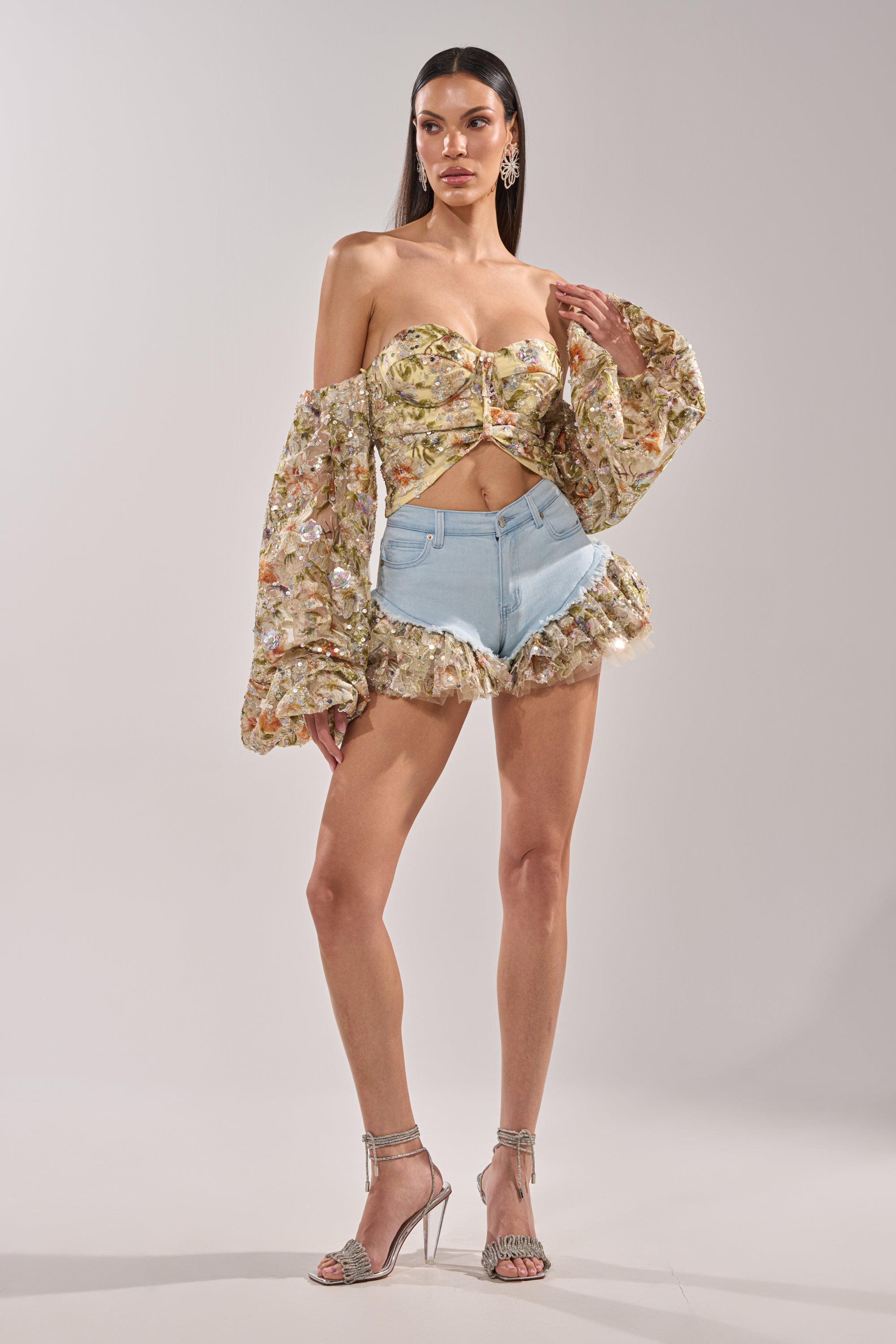 A woman poses against a plain background in the QUEEN OF IT ALL FLORAL DENIM SHORT, paired with a floral off-shoulder crop top featuring puffy sleeves, tulle ruffle trim, and silver high-heeled sandals. 6
