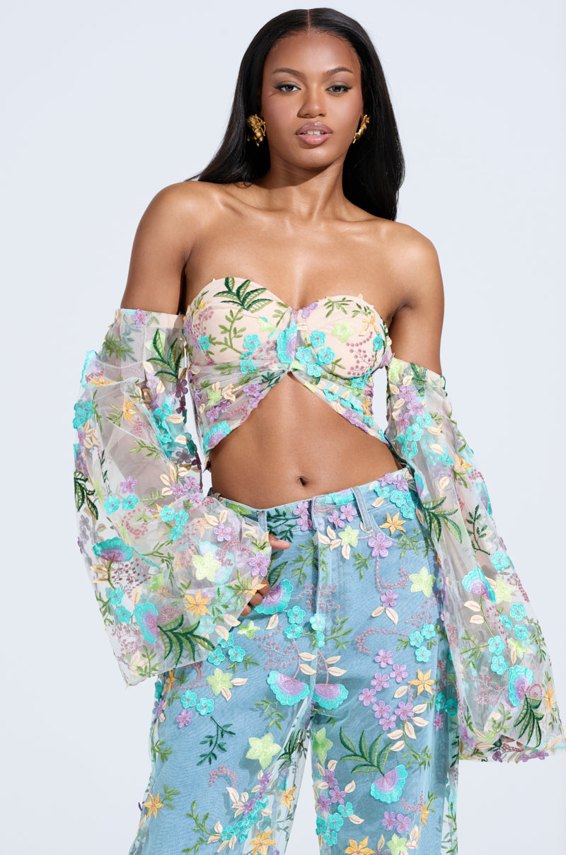 In the serene light blue setting, a woman captivates in the GARDENIA FLORAL EMBROIDERY TULLE TOP with transparent bell sleeves and matching high-waisted pants. Her cascading hair is adorned with shimmering gold earrings, enhancing her graceful ensemble.