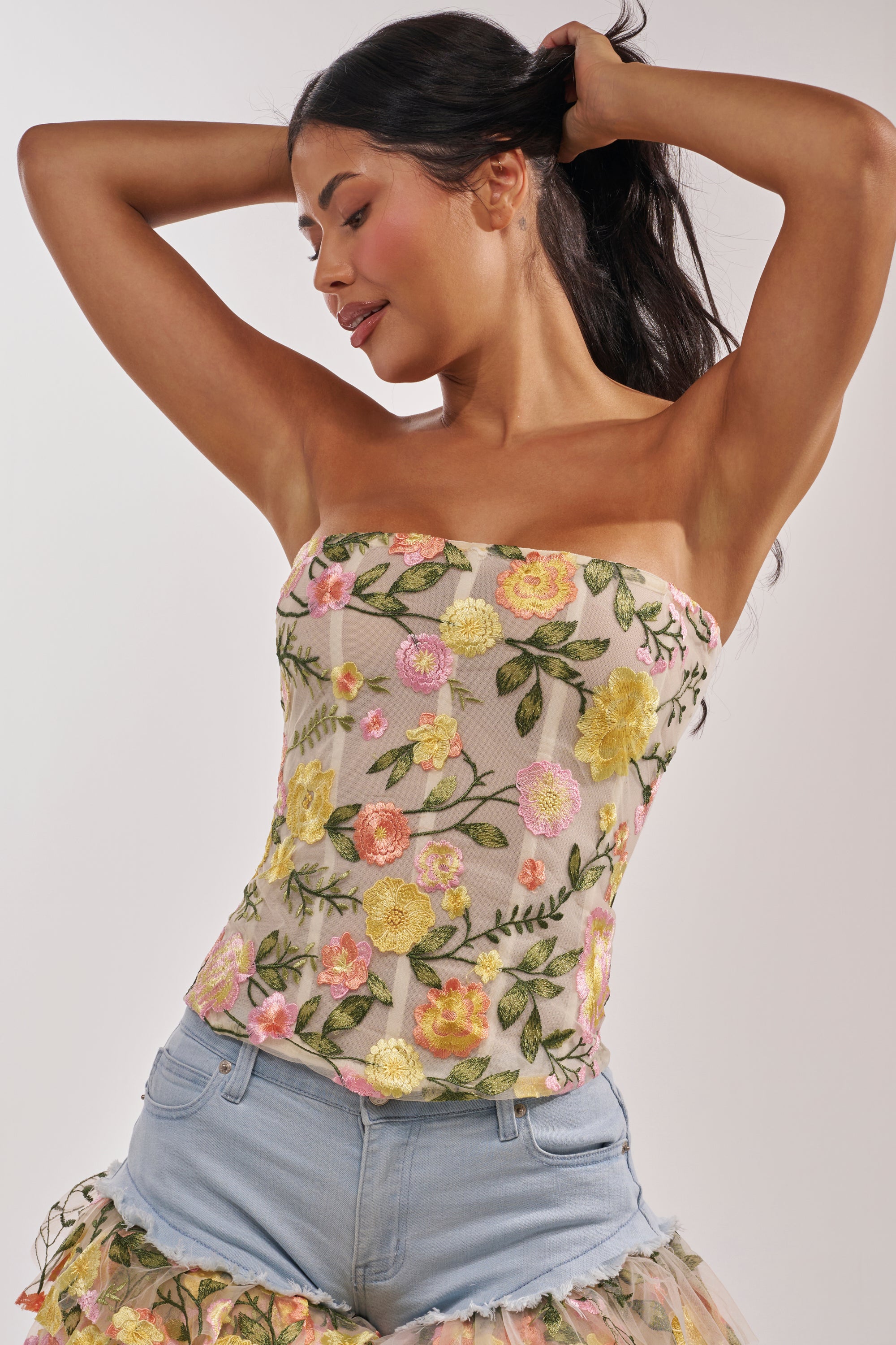 A woman smiles softly and poses with raised arms, wearing the VIVIAN FLORAL CORSET—a romantic strapless top with colorful floral embroidery—paired with light blue denim shorts. Her long dark hair is pulled back as she looks to the side. 1