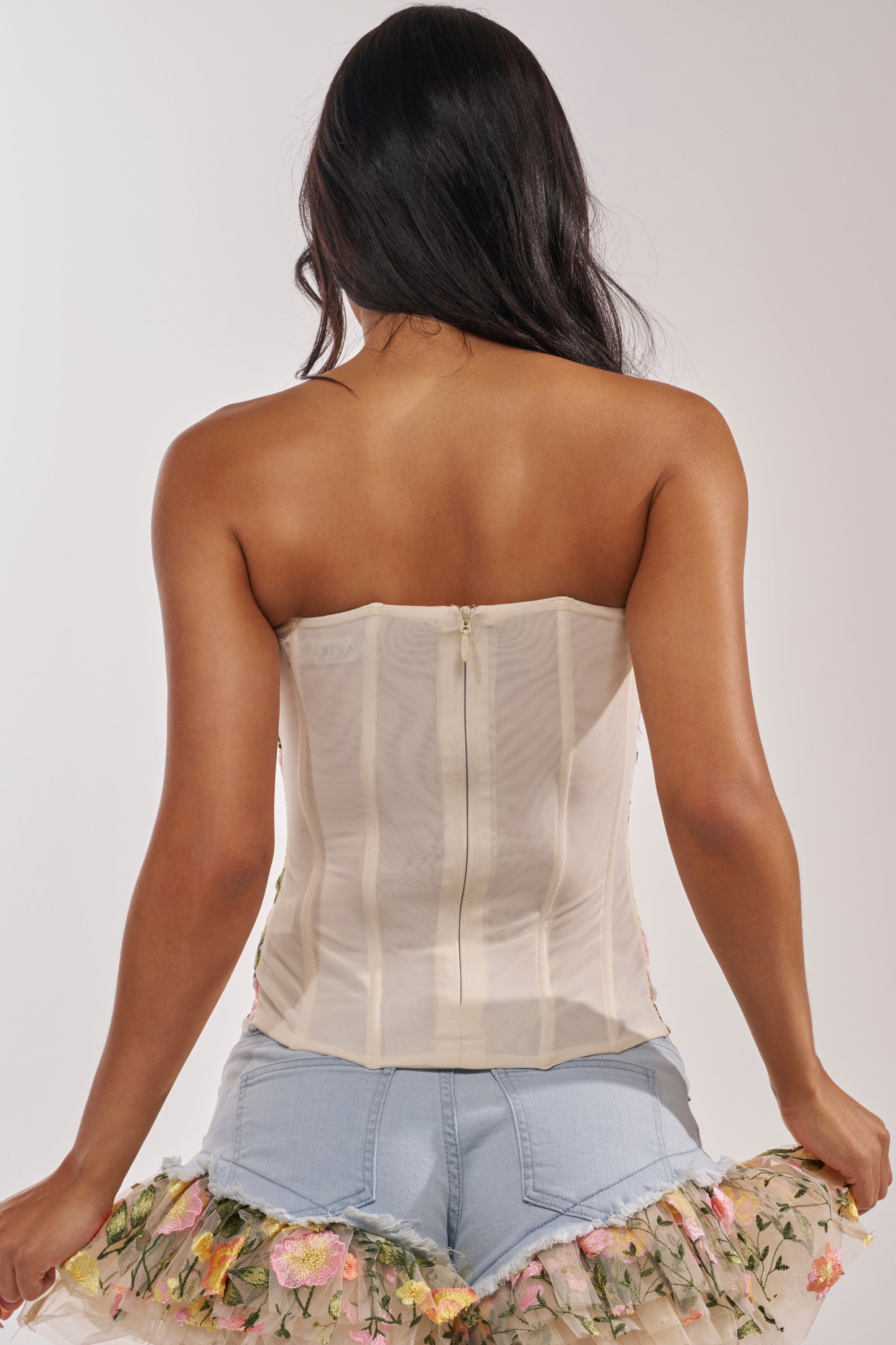 A woman with long dark hair is shown from behind, wearing the VIVIAN FLORAL CORSET with a visible back zipper and light blue shorts featuring floral embroidery and delicate tulle details. 3