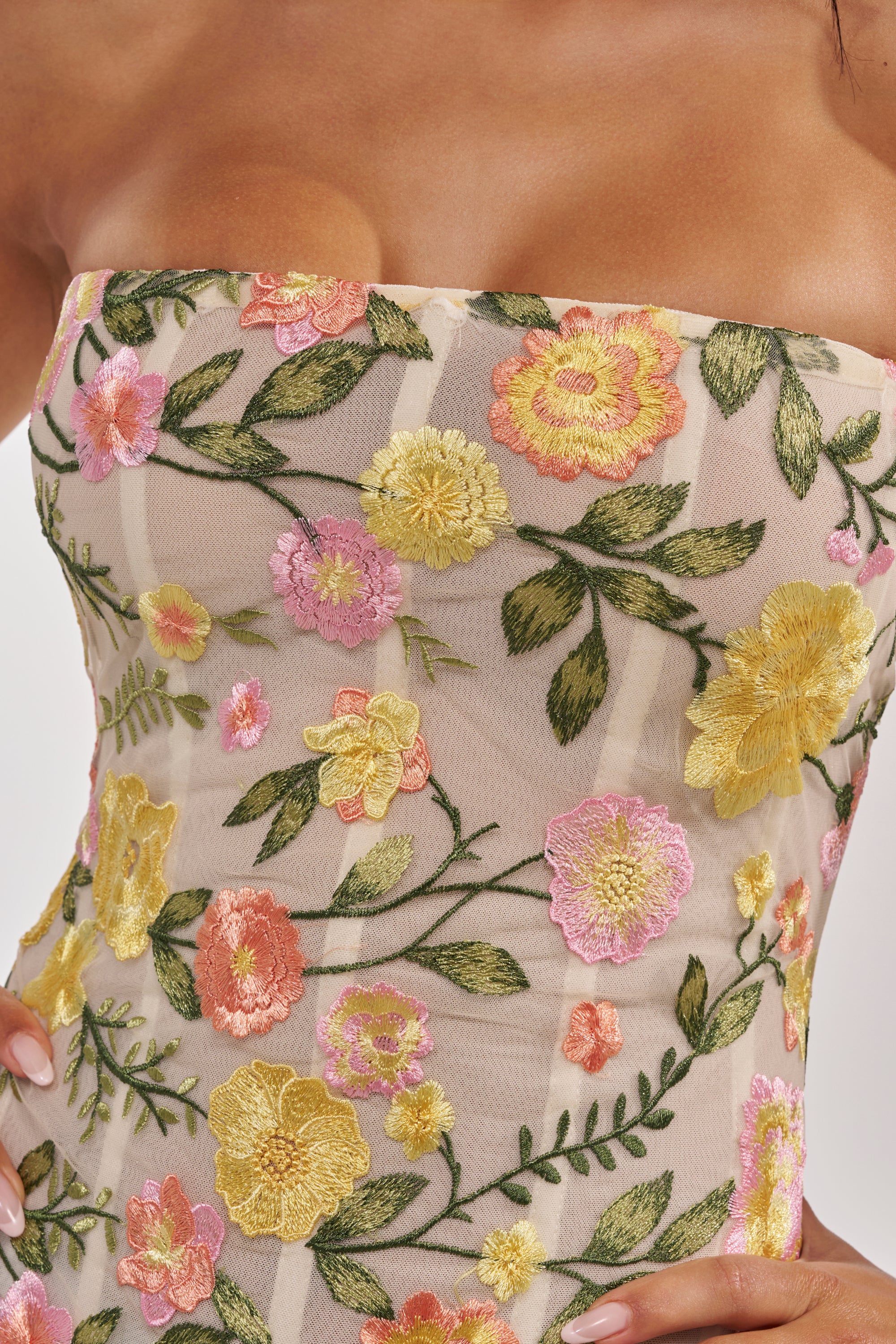 A close-up of the VIVIAN FLORAL CORSET, a strapless dress with yellow, pink, and green floral embroidery on sheer beige fabric. The hand-on-hip pose highlights its elegant corset silhouette. 7