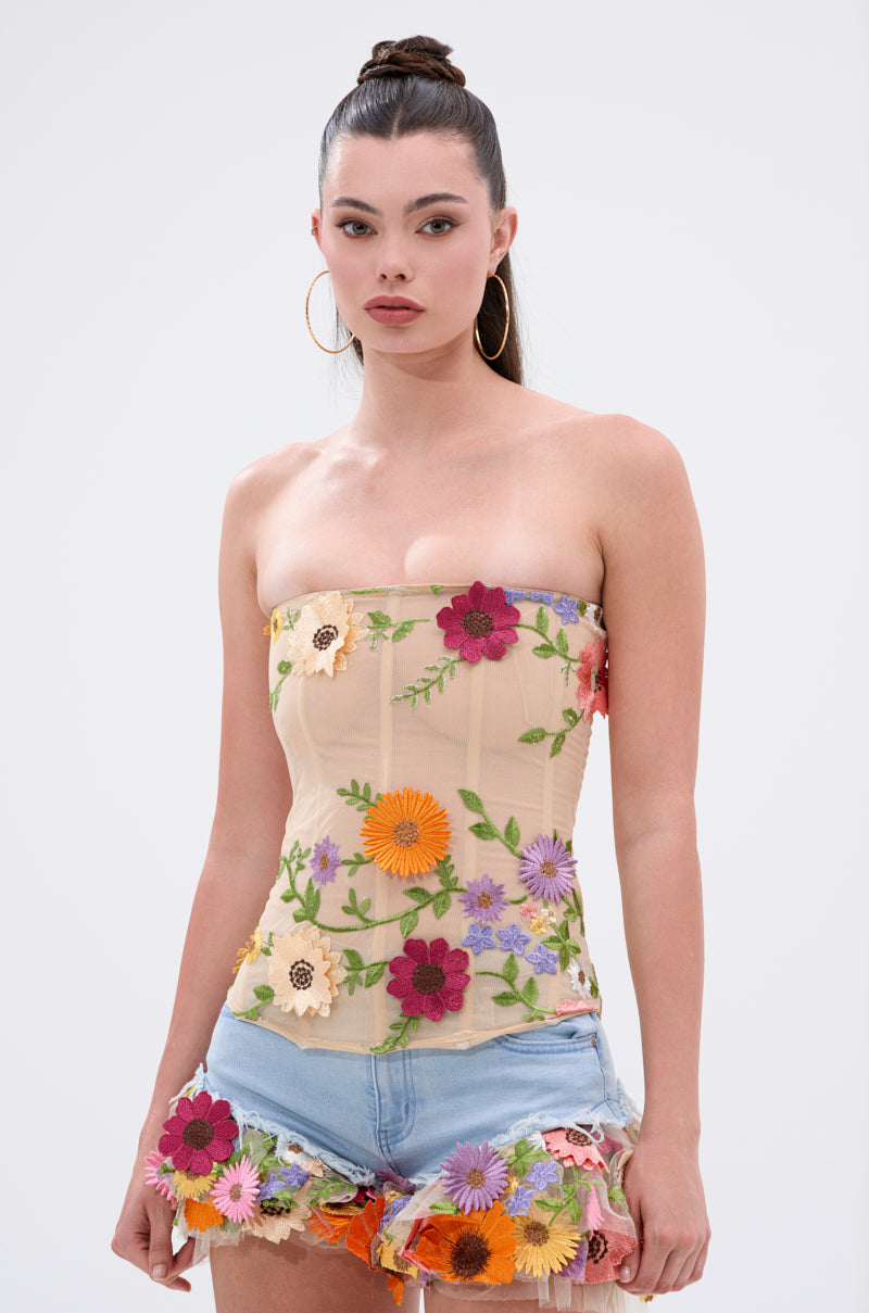 A woman with long dark hair in a high bun wears the VIVI FLORAL CORSET, featuring colorful 3D floral appliqués, paired with denim shorts. She stands against a plain light background.