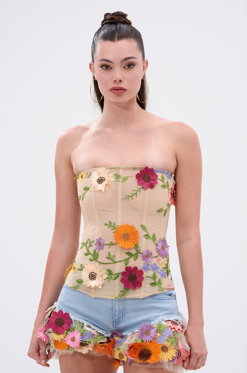 A woman wears the VIVI FLORAL CORSET, a strapless sheer mesh top with colorful floral appliques, paired with light blue denim shorts featuring matching flowers on the hem. She completes the look with hoop earrings and a top bun.